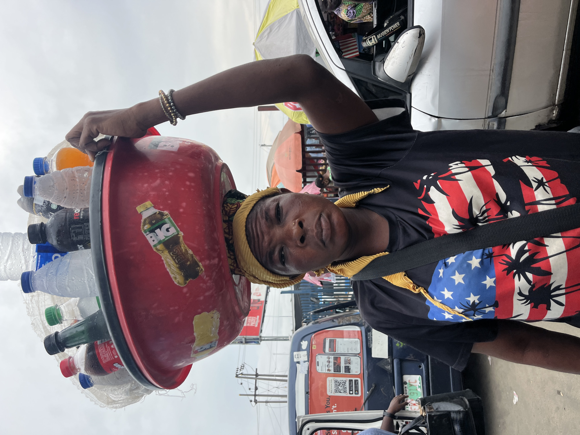 A street vendor in Lekki, Nigeria, balances a red bowl on her head filled with various bottled beverages. She wears a black t-shirt with a palm tree and American flag print, a yellow headscarf, and bracelets. The backdrop includes a commercial vehicle, street umbrellas, and the urban environment of Lekki.