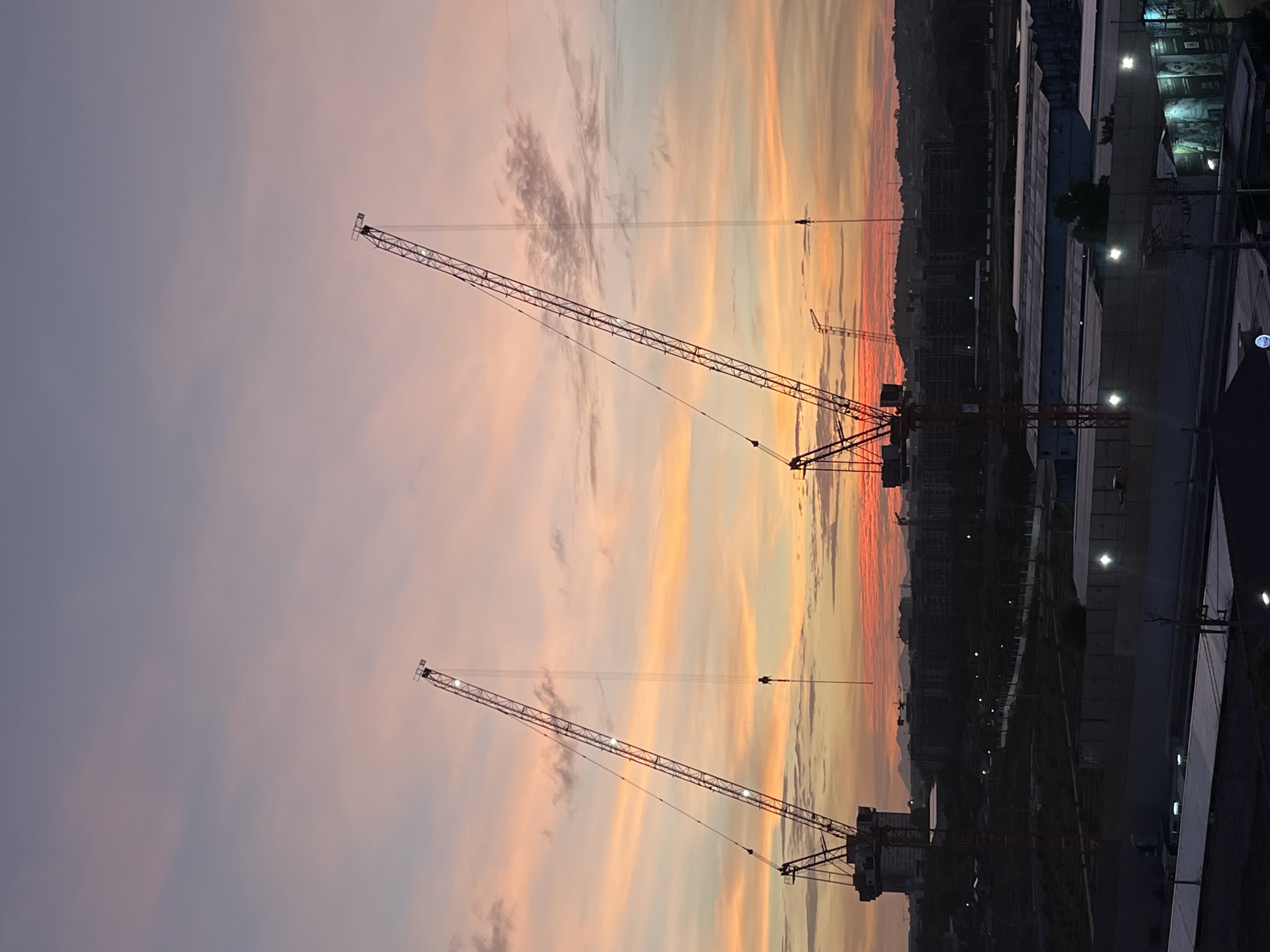 The image depicts a construction site in Pattaya City, Thailand, at sunset. Three cranes are silhouetted against the colorful sky, ranging from shades of purple to orange. The city skyline is visible in the background. To the right of the cranes, there appear to be industrial buildings or warehouses with lights.