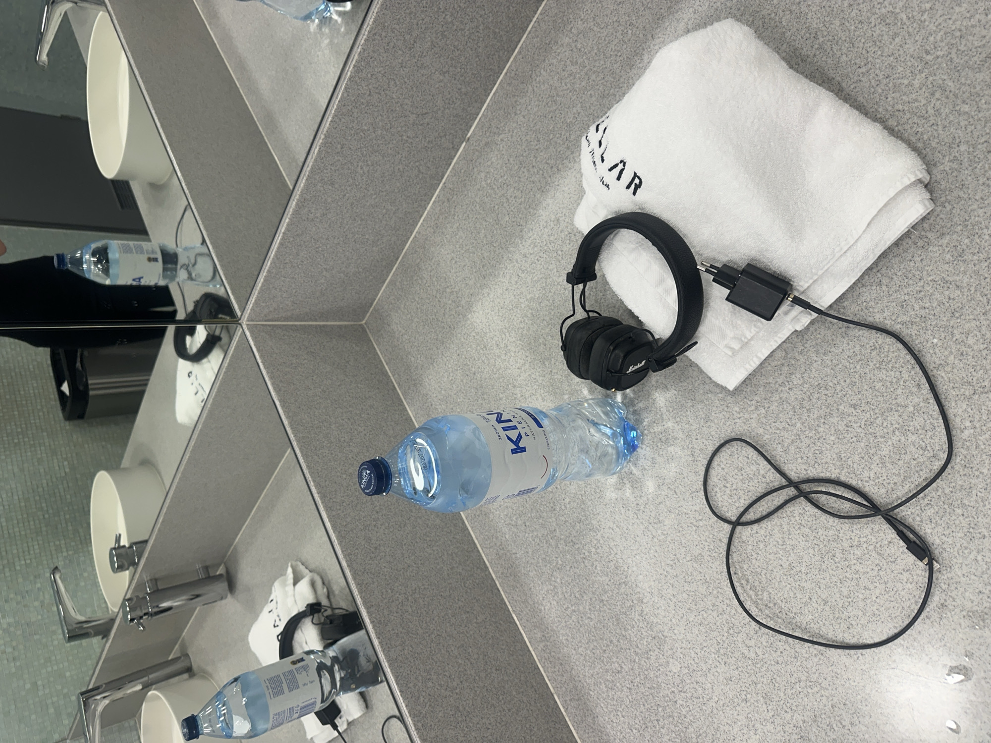 The image depicts a bathroom countertop in Unknown, Unknown, covered with various items. A folded white towel with black lettering rests near a pair of black Marshall headphones connected to a charging cable and adapter. A bottle of water, labeled "KINIA" and filled with water, lies nearby. The countertop is part of a mirrored installation, reflecting the same scene repeatedly with slight variations. Two white, cylindrical sinks with silver faucets are visible in the reflection. The wall behind the sinks is covered in a mosaic of pale green tiles. The countertop surface is a speckled gray material. The scene appears still and unoccupied.