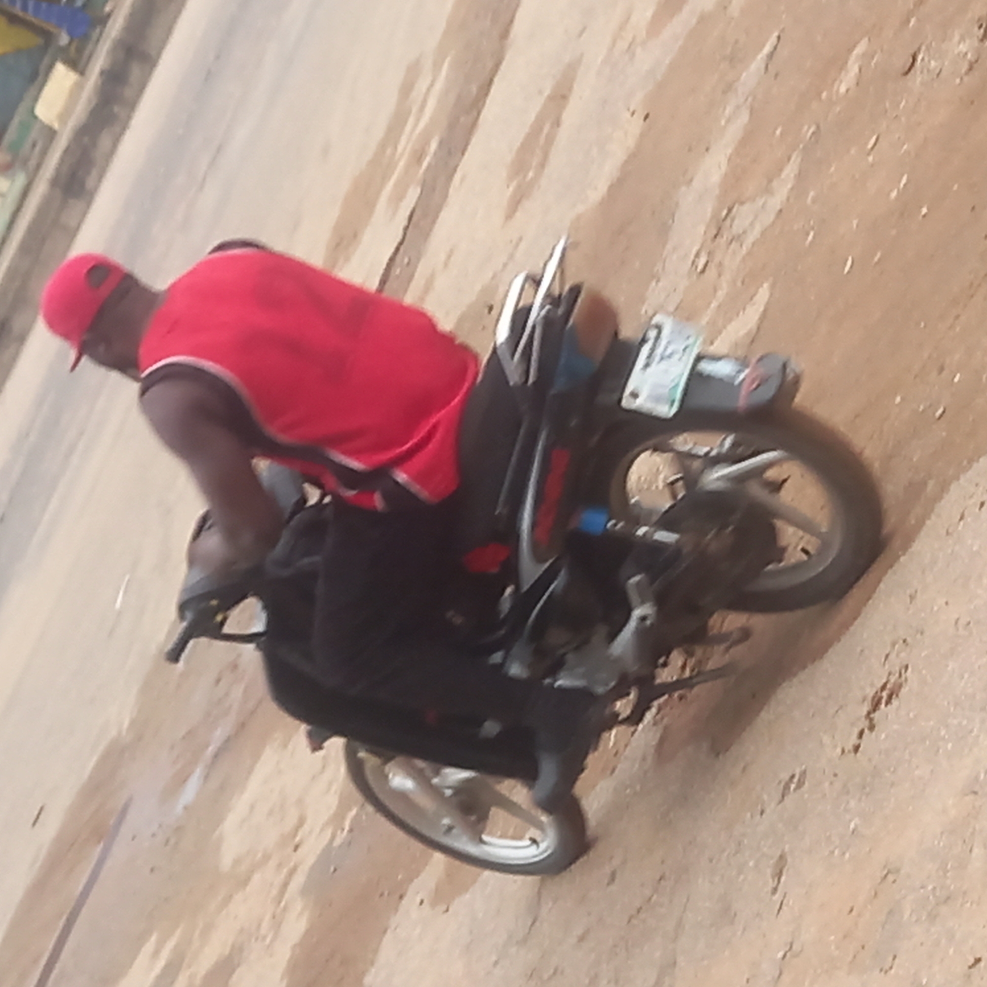An eye-level shot captures a person riding a motorcycle on a light brown, possibly dusty or unpaved road, with some lighter gray patches indicating mixed road surfaces. The image is tilted, suggesting a dynamic angle or movement.
The primary subject is a male individual, seen from behind and slightly to the left, wearing a bright red baseball cap and a red sleeveless top with white trim on the shoulders. His skin appears dark. He is seated on a black and grey motorcycle, which has a visible rear wheel, a silver-colored exhaust pipe, and a partially obscured license plate. The license plate appears to have some indistinct white characters on a dark background, with the number "1" faintly discernible. The front wheel of the motorcycle is also visible.
The background on the left is indistinct, showing faint shapes that could be distant buildings or foliage. The overall lighting suggests it is daytime. No specific time of day or weather conditions are explicitly clear, though the dusty road surface hints at dry conditions. The image does not convey any specific emotions. The location context provided is Sardauna, Nigeria.