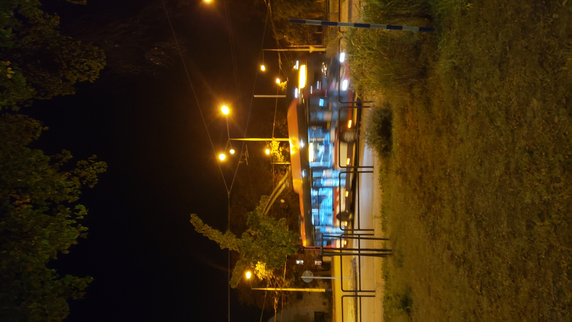 The image captures a night scene in Prešov, Slovakia, featuring a blurred trolleybus in motion on a street. The trolleybus is orange and white with illuminated windows. In the foreground, there is a grassy area with some bushes and a black metal fence. In the background, streetlights cast a warm glow on the dark sky, and trees with foliage are visible. The overall impression is a typical urban environment at night, with public transportation operating.