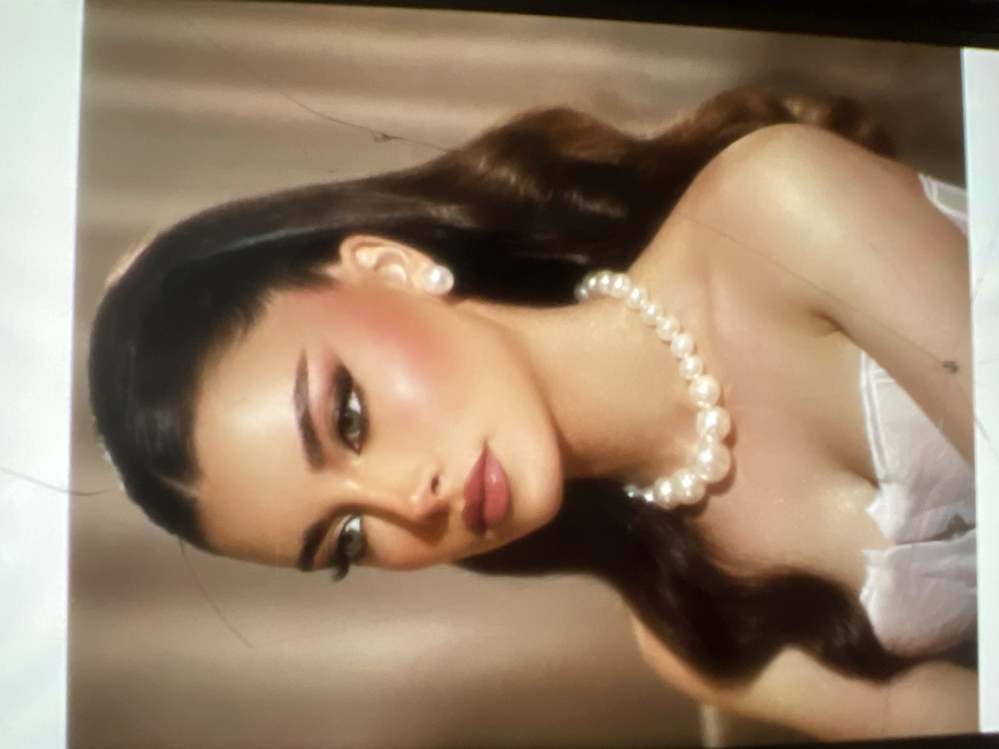 This is a medium shot portrait of a young woman with dark hair styled in waves and a high ponytail. She is wearing a pearl necklace and pearl earrings, and her makeup includes dramatic winged eyeliner and a soft pink lip. She is dressed in a white, strapless garment. The background is a neutral, textured curtain. There is no discernible location in Kara-Dıykan, Kyrgyzstan.