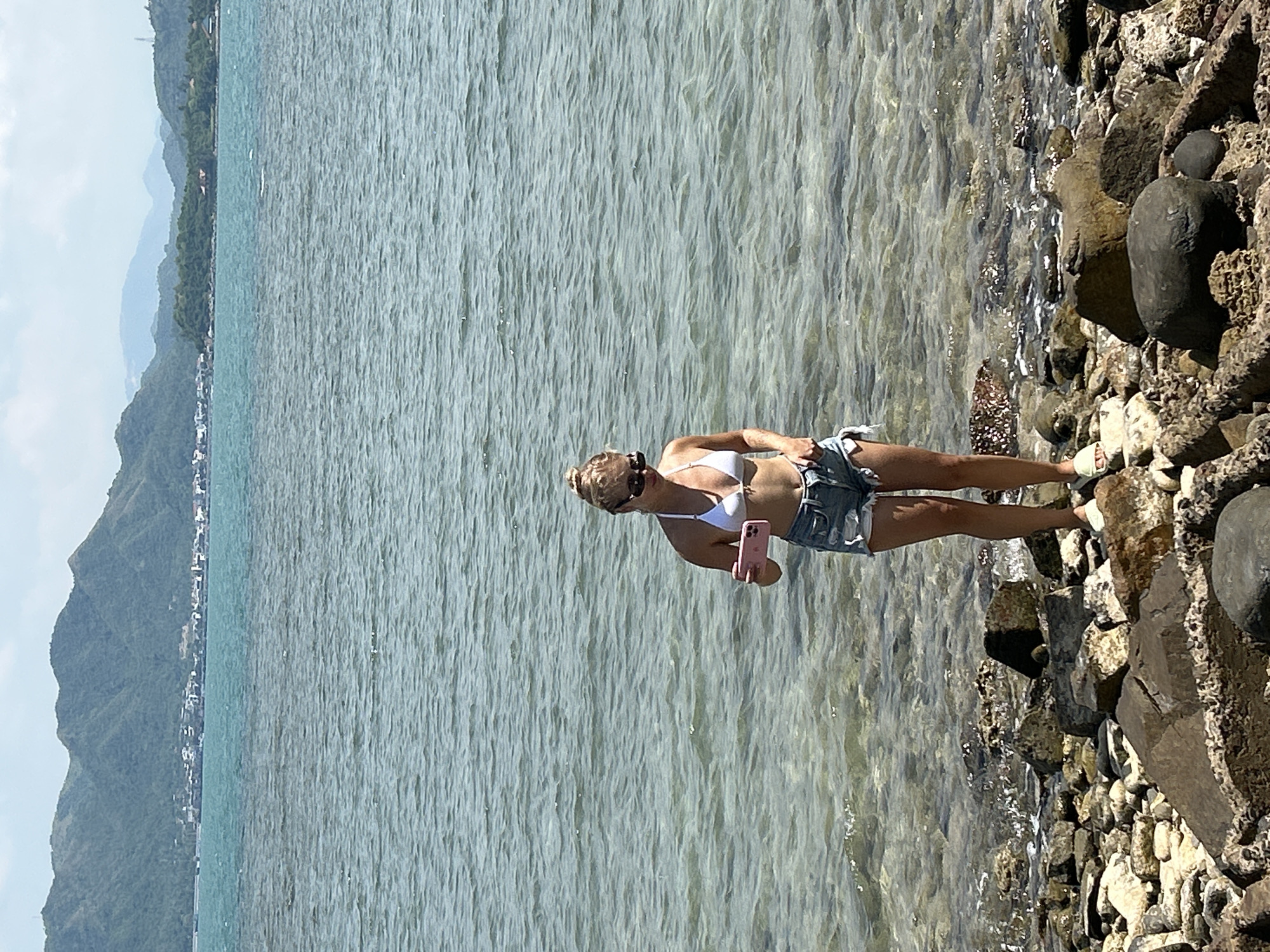 A young woman stands on a rocky shore at the edge of the sea. She is wearing a white bikini top and denim shorts. She is holding a pink smartphone in her right hand, and her left hand is in her pocket. She is looking down at the phone. Her hair is blonde and pulled up in a bun. She is wearing sunglasses. The sea is calm and clear, with visible rocks beneath the surface. In the background, there are green mountains and a distant coastline with buildings. The sky is partly cloudy. The image appears to have been taken in Ninh Ích, Vietnam, given the landscape.