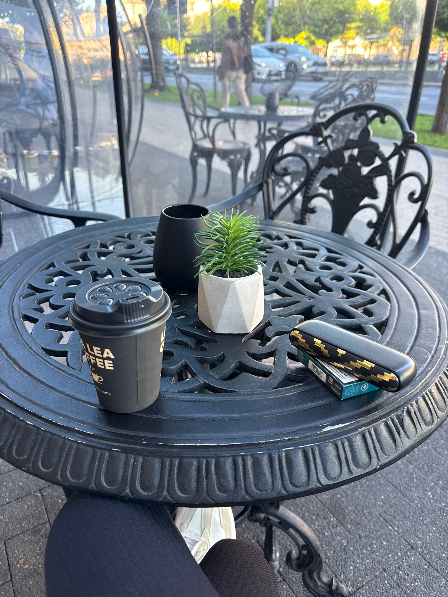 An outdoor cafe scene in Chișinău, Moldova, features a black metal table with ornate detailing. On the table are a black coffee cup with "ALEA COFFEE" printed on it, a small potted plant with green foliage in a concrete planter, and a black and gold electronic smoking device resting on a blue box of tobacco sticks. In the background, blurred figures are visible through a clear enclosure, and the street with cars and trees can be seen. The foreground shows a person's legs wearing black leggings and white sneakers.