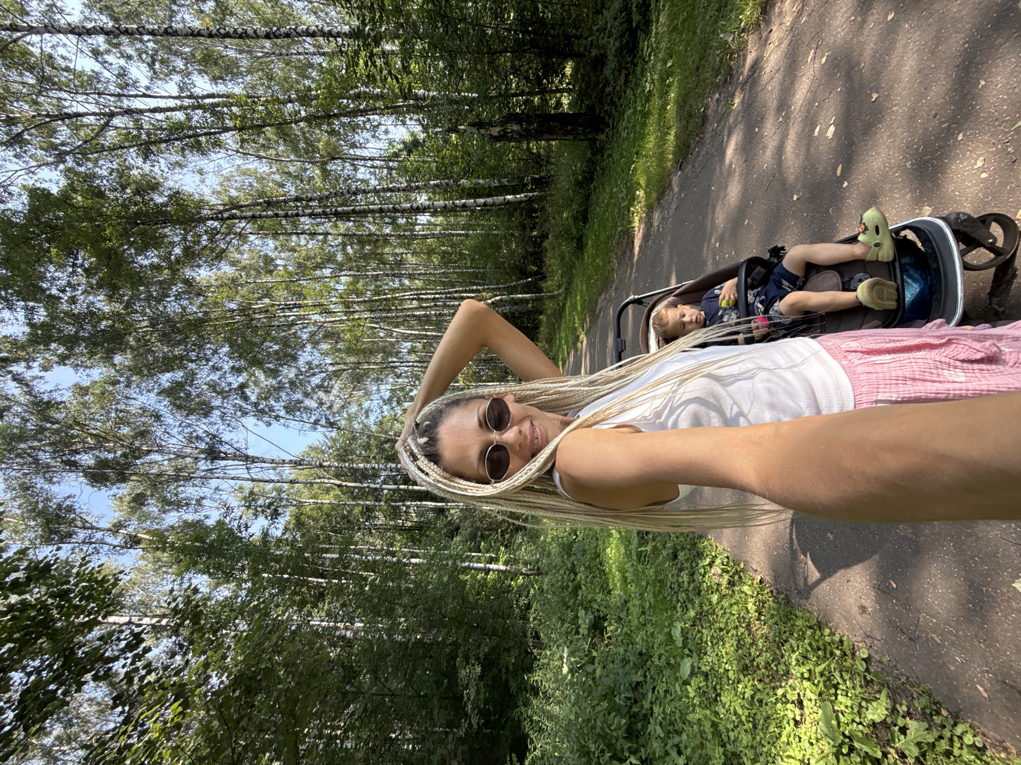 In the city of Dumino, Russia, on a paved path through a birch forest, a woman with blonde braided hair and sunglasses is taking a selfie. She is wearing a white tank top and a pink skirt. Next to her, a toddler sits in a stroller, wearing a dark blue t-shirt, dark blue shorts, and light green Crocs. The child is holding a yellow ball. Sunlight filters through the trees, creating dappled shadows on the path and surrounding greenery.