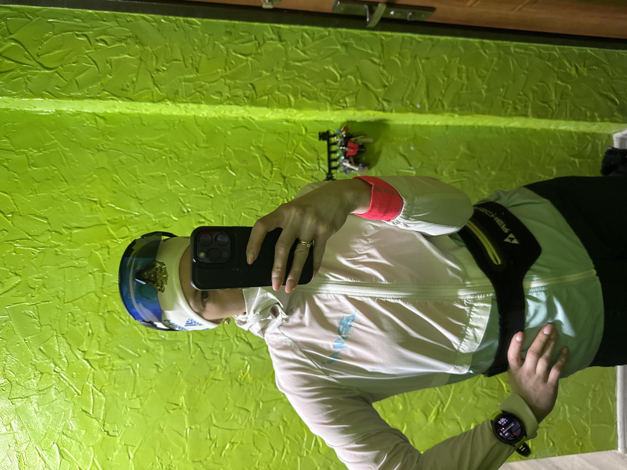 A person stands indoors, taking a selfie. They are wearing sports attire consisting of a white zip-up jacket with light blue accents, a black belt with yellow trim around their waist, a white headband, and blue goggles resting on their head. A smartwatch is visible on their left wrist. They hold a black smartphone in their right hand, taking the photo. The background shows a textured green wall, and a section of a wooden door with a metal latch is visible near the top of the frame. The location is Chaikovsky, Russia.