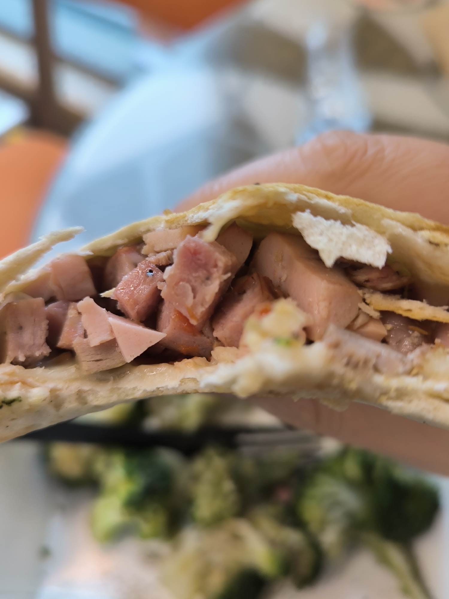 A close-up, slightly blurry shot shows a person's hand holding a halved quesadilla. The quesadilla's filling, consisting of diced ham and some leafy greens, is clearly visible. In the background, out of focus, is a plate of steamed broccoli. The setting appears to be indoors, possibly a restaurant or home kitchen, with soft lighting. There are no discernible landmarks or indicators to suggest a specific location within Novosibirsk, Russia.