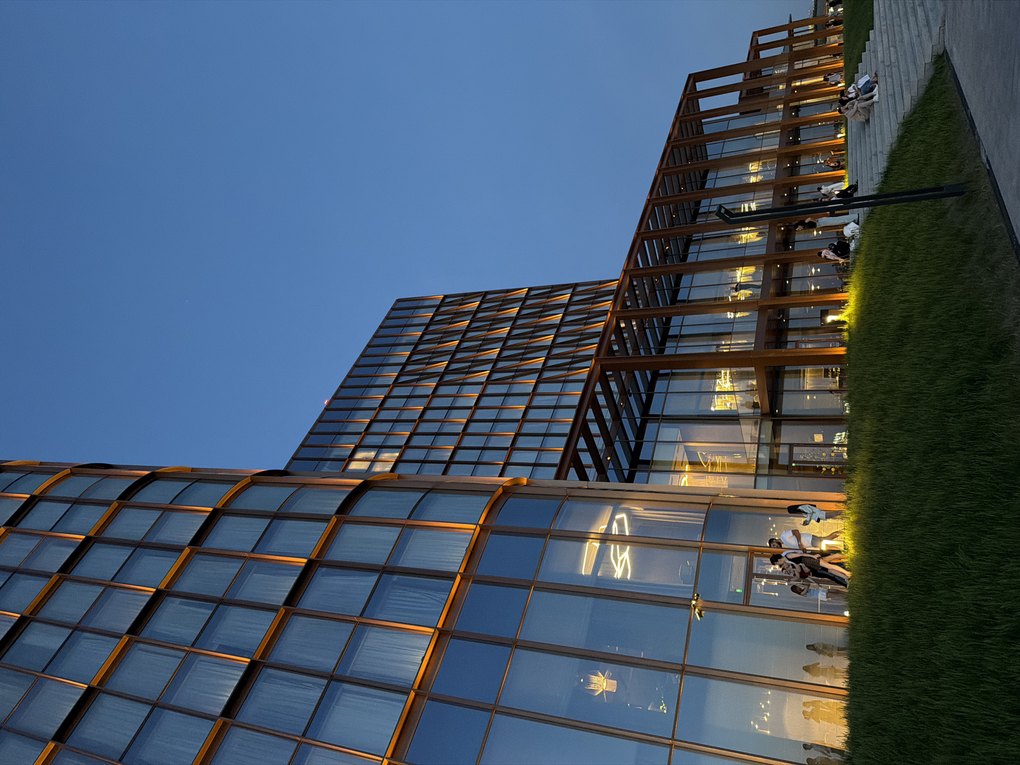 The image presents an architectural scene, likely at twilight, in an urban environment. Several modern buildings dominate the view, characterized by their glass facades and geometric structures. One building features a curved glass exterior with a grid-like pattern, while another adjacent building has a more angular design with a brown metal framework.
On the right side of the frame, people are seated or standing on a set of stairs or tiered platforms. These individuals are illuminated by warm lights embedded in the ground, which also highlight the adjacent grassy area. A dark, pole-like object is also present.
The sky is a clear, deep blue, indicating the time of day. The reflective surfaces of the buildings capture the ambient light, creating highlights and shadows. The overall composition is dynamic, with converging lines and contrasting textures.