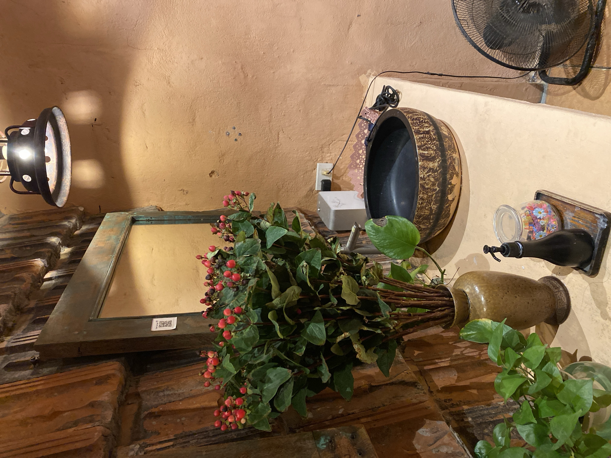 This is a medium shot, eye-level, portrait-oriented photo taken indoors. The scene is a room with warm, terracotta-colored walls. In the foreground, a bouquet of greenery with red berries is arranged in a textured, golden vase. To the left of the bouquet, a vintage-style mirror with a weathered, greenish-brown frame rests against a brick surface. Above the mirror, a black industrial-style lamp with a visible lightbulb casts shadows on the wall.
Further into the background, a decorative, dark basin with a patterned rim is mounted on the wall, possibly a sink or a vessel. To the right of this basin, a black oscillating fan is visible against the wall. On a ledge beneath the basin, a small bottle with a spout, possibly a soap dispenser, is mounted. There is also a clear container filled with colorful, mosaic-like objects, and a white electrical outlet and switch are visible on the wall.
The lighting suggests it could be daytime, with artificial light from the lamp also contributing. The overall atmosphere feels rustic and eclectic, with a mix of natural elements and vintage decor. There is no visible text except for a small QR code sticker on the mirror. The location context provided is Da Nang, Vietnam.