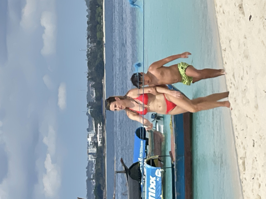 An outdoor, eye-level photograph depicts a woman and a child walking on a sandy beach in Zanzibar, Tanzania, under a partly cloudy sky. The woman, appearing to be an adult, wears a red two-piece bikini and has dark hair. She walks barefoot on the light-colored sand, looking straight ahead. To her right and slightly behind her, a young male child, shirtless and wearing green patterned swim trunks and dark-rimmed goggles on his head, walks barefoot. A blue rope or line extends horizontally across the frame at their mid-torso height. In the clear, shallow, turquoise water behind them, several boats are visible. One boat features a blue canopy with the partial text "Nixx" in white. In the distant background, across deeper blue water, a coastline with buildings and green vegetation is visible under the bright daytime sun.