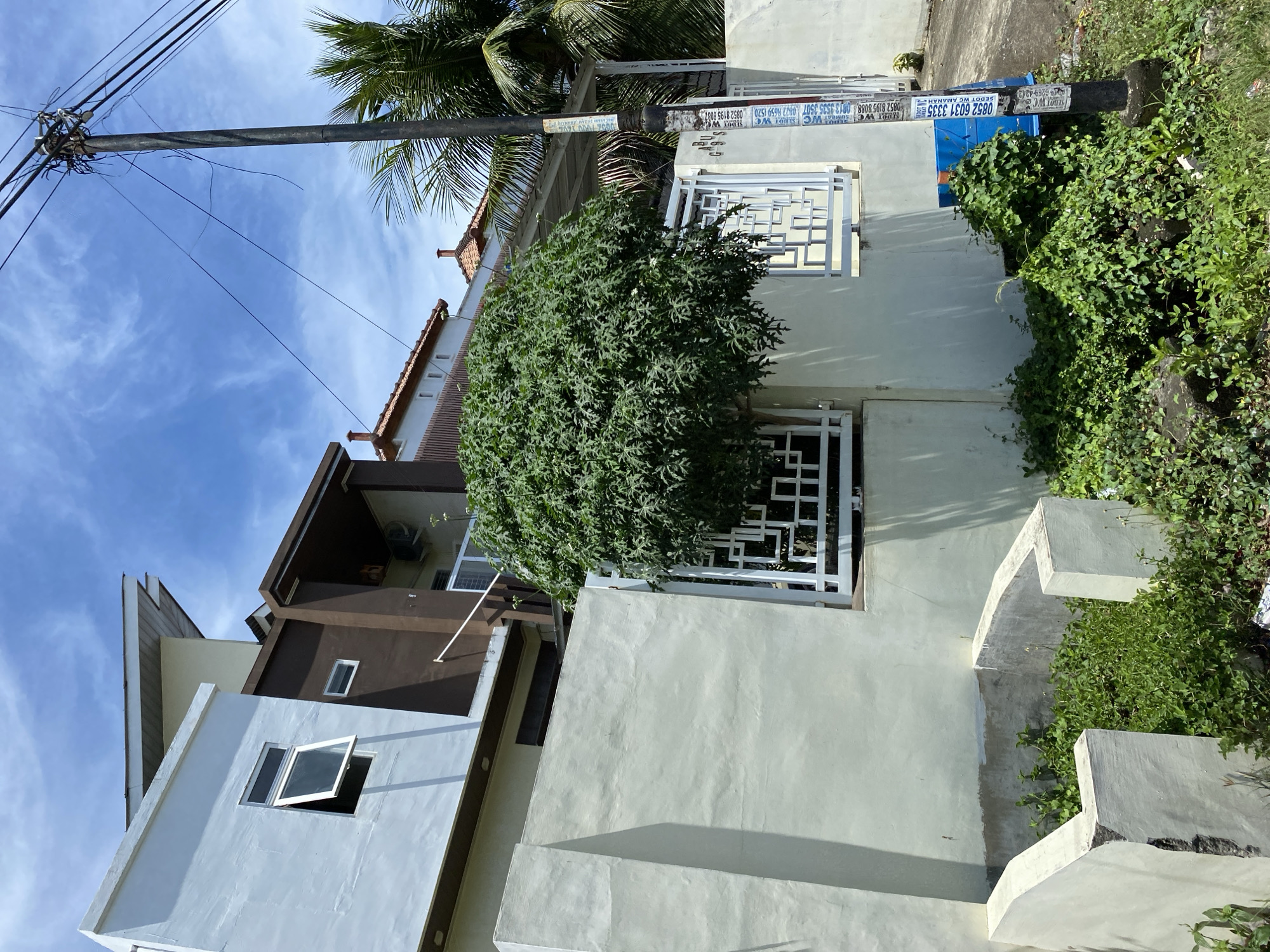 The image shows a street scene in Banda Aceh, Indonesia, on a clear day with a blue sky and scattered clouds. The foreground is dominated by a section of a light-colored concrete wall and a small, overgrown planter box. A lush, green tree with dense foliage stands next to the wall, partially obscuring decorative white metal fencing.
Behind the wall, modern houses with multi-level architecture are visible. One house has a white exterior and a brown upper section, featuring various windows, including an open casement window. Another house behind it has a reddish-tiled roof. A utility pole with numerous wires stands to the right of the frame, adorned with several paper advertisements. A blue plastic bin is situated near the base of the utility pole, partially covered by vegetation. Palm trees are visible in the background, suggesting a tropical climate. The overall impression is one of a residential area in a developing city.