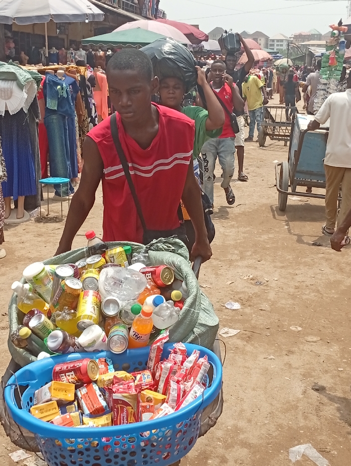 The image depicts a crowded outdoor market scene in Fegge, Nigeria, during daylight hours. In the foreground, a young male wearing a red sleeveless shirt with white horizontal stripes and a dark shoulder bag pushes a two-wheeled cart. The cart carries two distinct loads: on top, within a green woven sack, is an assortment of discarded aluminum beverage cans and plastic bottles, some containing residual liquids. Beneath this, in a blue plastic laundry basket, are various packaged snack foods, primarily biscuits and instant noodles.
Behind the foreground subject, multiple individuals navigate the market. One person directly behind him also carries a large black bag on their head. Further into the background, other people are visible, including a male in a red shirt talking on a mobile phone and another person pushing a different two-wheeled cart. Market stalls line the area, displaying clothing on mannequins, with various colored umbrellas and awnings providing shade. The ground is unpaved, consisting of dirt, with some scattered debris. Buildings are visible in the far background under a bright sky.