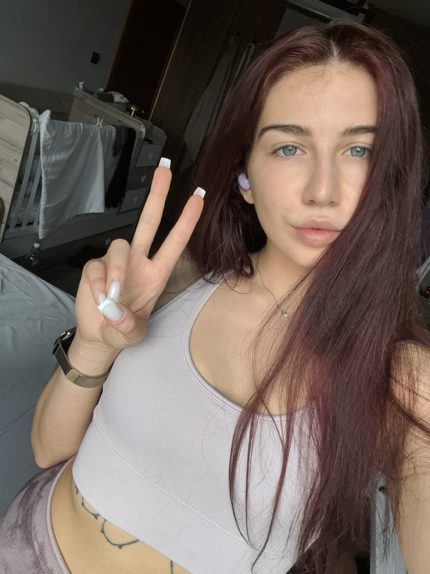 A young woman with long reddish-brown hair and striking light blue eyes takes a selfie in what appears to be an indoor setting, likely a bedroom, in Stara Zagora, Bulgaria. She is looking directly at the camera with a neutral expression, showing a slight hint of a smile. Her left hand is raised, making a peace sign, prominently displaying her long, square-tipped nails with white French tips.
She is wearing a light lavender or pale purple ribbed sports bra or crop top, and what appears to be patterned leggings are visible at the bottom left of the frame. A delicate silver-toned necklace with a small pendant rests on her collarbone, and a gold-toned mesh watch adorns her left wrist. A tattoo, resembling a circular or floral pattern, is visible on her left hip/lower abdomen. She has a single light purple wireless earbud in her right ear.
The background elements suggest a lived-in room. To her left (viewer's right), a light-colored wooden crib or a bed with white fabric draped over it is visible. Behind it, a wooden dresser or chest of drawers can be seen, partially obscured, with a few items on top. Further back, a darker wall or door is present. The lighting is soft and natural, suggesting it is daytime. No visible text is present in the image.