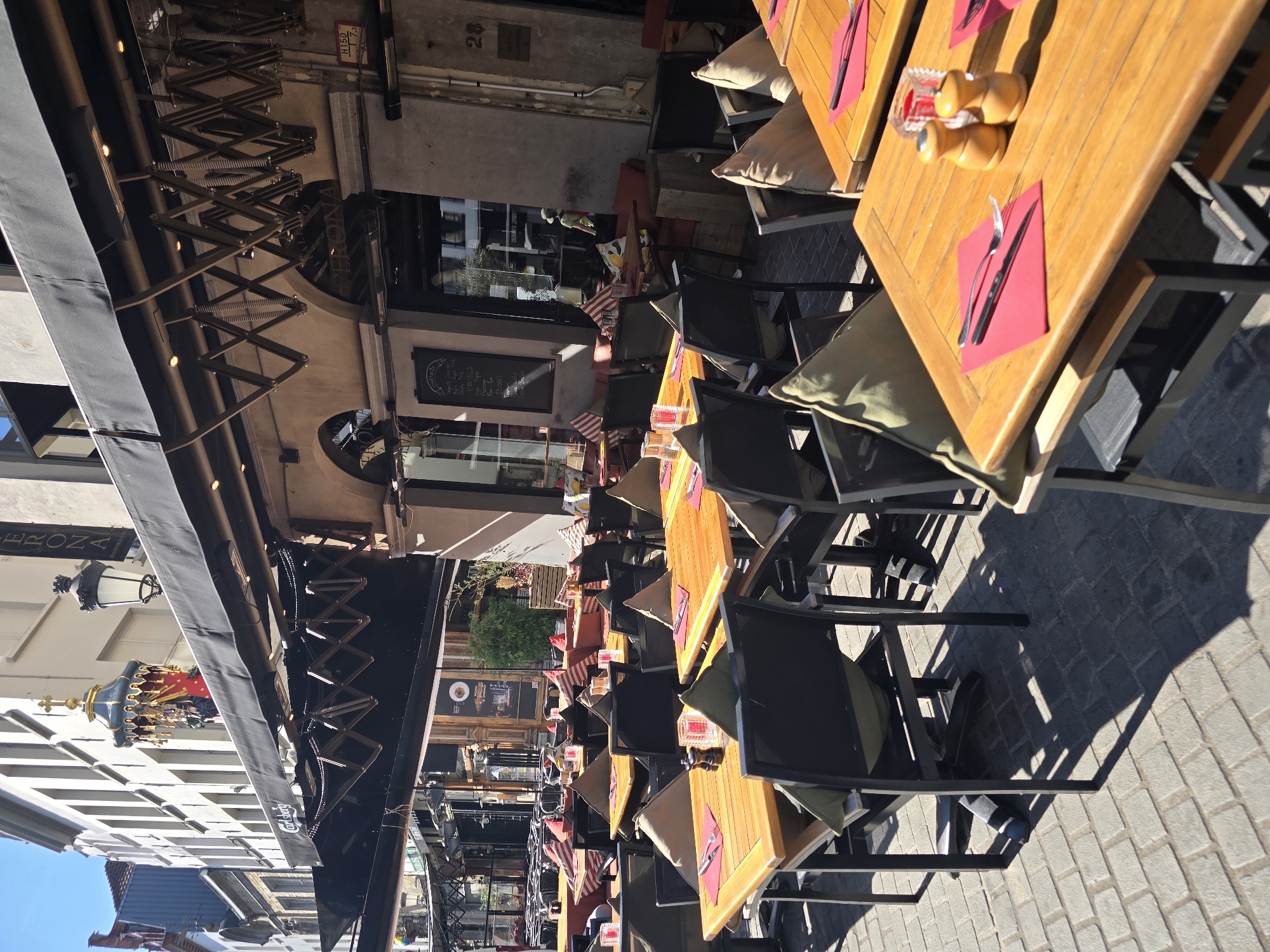 This image depicts the outdoor seating area of a restaurant located on the Meir, a prominent shopping street in Antwerp, Belgium. The scene is bathed in bright sunlight, casting distinct shadows on the cobblestone pavement.
In the foreground, several wooden tables are set with red napkins, cutlery, and salt and pepper shakers, suggesting that the establishment is open for business or preparing to serve customers. The tables are surrounded by chairs with cushions, some of which are a light green or beige color, and others are red and white striped.
The restaurant itself appears to be multi-story, with a large black awning extending over the outdoor seating area. Behind the awning, the facade of the building is visible, featuring arched windows and signage that includes "VERONA RISTORANTE". A decorative element, possibly a weather vane or a historical sculpture, is perched on top of a building in the background.
To the right of the building, a number "28" is visible on a plaque, further indicating a specific location on the street. A person wearing a light-colored shirt and shorts can be seen inside the restaurant, near the entrance.
The overall impression is one of a lively urban setting, with the architecture of Antwerp providing a backdrop to the dining experience.