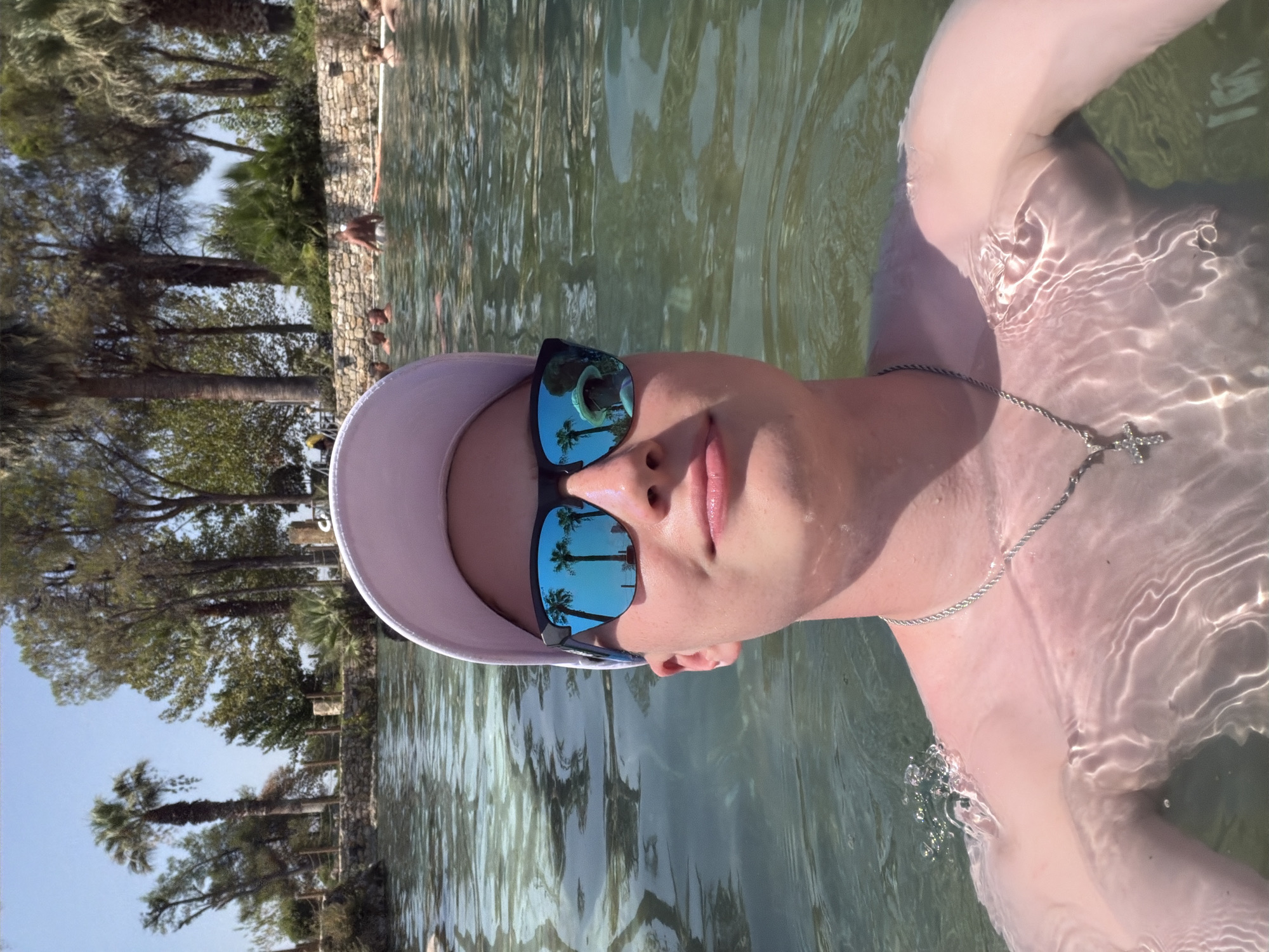 A young person is taking a selfie in a geothermal pool in Pamukkale, Turkey. The person is wearing a light-colored baseball cap and dark sunglasses, which reflect palm trees and the sky. They are partially submerged in clear, green water, with ripples from movement visible on their skin and shoulders. A silver chain with a cross pendant is visible around their neck. In the background, other people are relaxing in the water, and a stone wall with fencing and lush vegetation, including palm trees, lines the edge of the pool. The sky is clear and blue.
