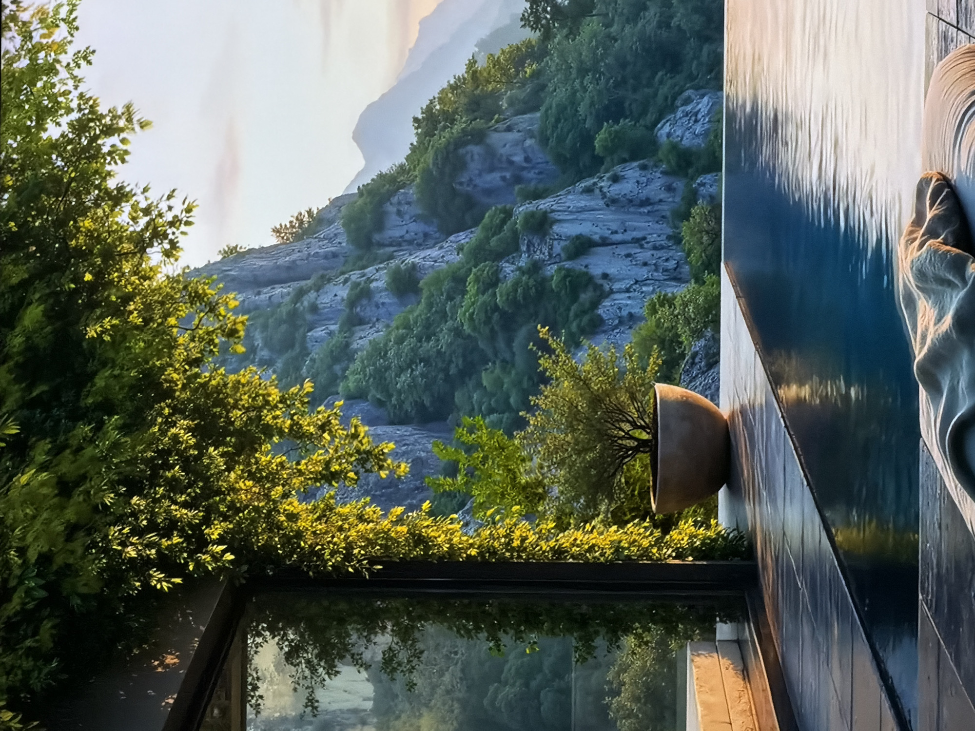 The image captures a serene outdoor scene, presented rotated 90 degrees counter-clockwise from its natural upright orientation. It depicts a luxurious architectural setting integrated with a vast natural landscape, likely a terrace or balcony overlooking mountains.
In the foreground (occupying the right side of the rotated image), a contemporary water feature is prominent. It consists of a dark tiled wall with water cascading down, creating a reflective, rippled surface. Resting on the edge beside the water is a light-colored, soft fabric, possibly a towel or part of an outdoor furnishing. Below this water feature, a large, rustic, terracotta-colored planter holds a small, verdant tree or shrub. Further down, a dark, reflective surface, likely a window or a lower pool, mirrors the surrounding foliage and the distant terrain.
The midground and background (filling the left side of the rotated image) showcase a majestic natural environment. Dominating this area is a rugged mountain range or large hillside characterized by grey, rocky outcrops and dense patches of vibrant green trees and shrubs. The lush foliage is illuminated by soft, warm sunlight, suggesting either early morning or late afternoon. The distant mountain peaks appear somewhat hazy against a bright sky, emphasizing their scale and the depth of the landscape.
No people are visible, and no specific activity or event is taking place. The overall mood is tranquil and luxurious, emphasizing natural beauty and sophisticated design. The warm, diffused lighting contributes to a peaceful atmosphere. No text is visible in the image. While the location context provided is Sofiivska Borshchahivka, Ukraine, the prominent, rugged mountainous terrain depicted suggests a naturally dramatic landscape, which may indicate a unique property or a broader regional view.