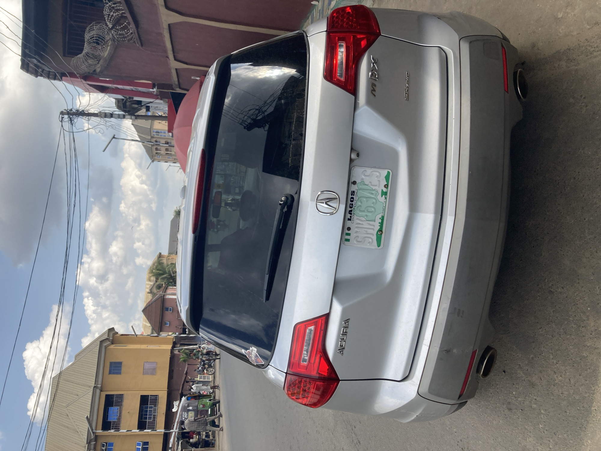 A silver Acura MDX is parked on the street in Umuode, Nigeria. The rear of the vehicle is prominently displayed, revealing a Lagos license plate. The surrounding environment includes buildings, power lines, and other vehicles. People are visible in the background.