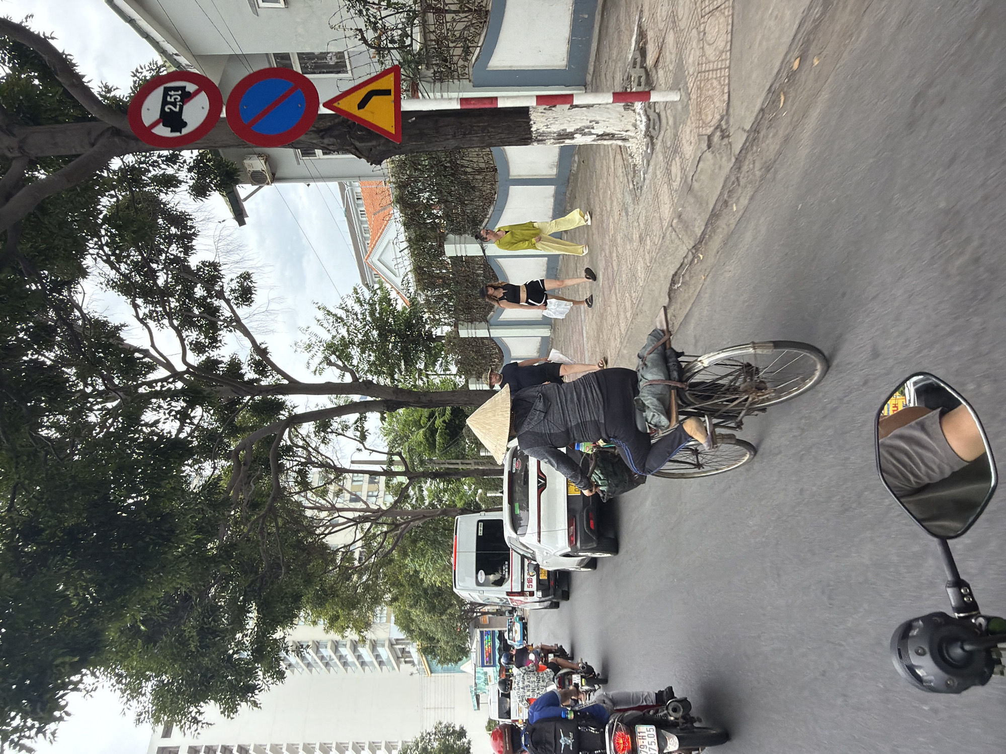 In this image, a view from the perspective of a motorbike rider in Nha Trang, Vietnam, reveals a busy street scene. A woman wearing a conical hat rides a bicycle in the foreground, her face obscured by the hat. She is carrying a bundle on the rear rack of her bicycle. Behind her, several motorbikes are also present in traffic.
To the right of the street, a sidewalk runs alongside a building. Three pedestrians are walking on the sidewalk: a woman in black shorts and a crop top, another woman in a yellow outfit, and a man in a dark t-shirt and shorts. A red and white striped pole with traffic signs is positioned next to the sidewalk. The signs include a circular sign with a red border and a truck symbol with "2.5t" inside a red circle and slash, indicating a weight restriction for trucks. Below that is a circular blue sign with a red border and a red diagonal bar, signifying "no stopping" or "no parking". A triangular yellow sign with a black arrow pointing left is also visible.
The background shows a high-rise building and trees lining the street. The road is paved, and traffic appears to be moderate. The overall atmosphere suggests a typical urban environment in Vietnam.
