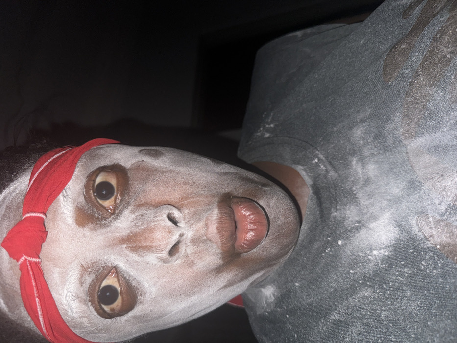 The image shows a close-up, front-facing view of a single individual located in Sabon Tasha, Nigeria. The subject, an adult with dark skin, has their face heavily coated with a white powdery substance, which extends onto their neck and a dark grey t-shirt. A red fabric headband is worn across the forehead. The subject's eyes are wide open, with dilated pupils and light brown sclera. The mouth is slightly open, with lips puckered forward, and the tip of the tongue is partially visible. The t-shirt is also dusted with the white powder. The background is dark and indistinct, indicating low-light conditions or an interior setting.