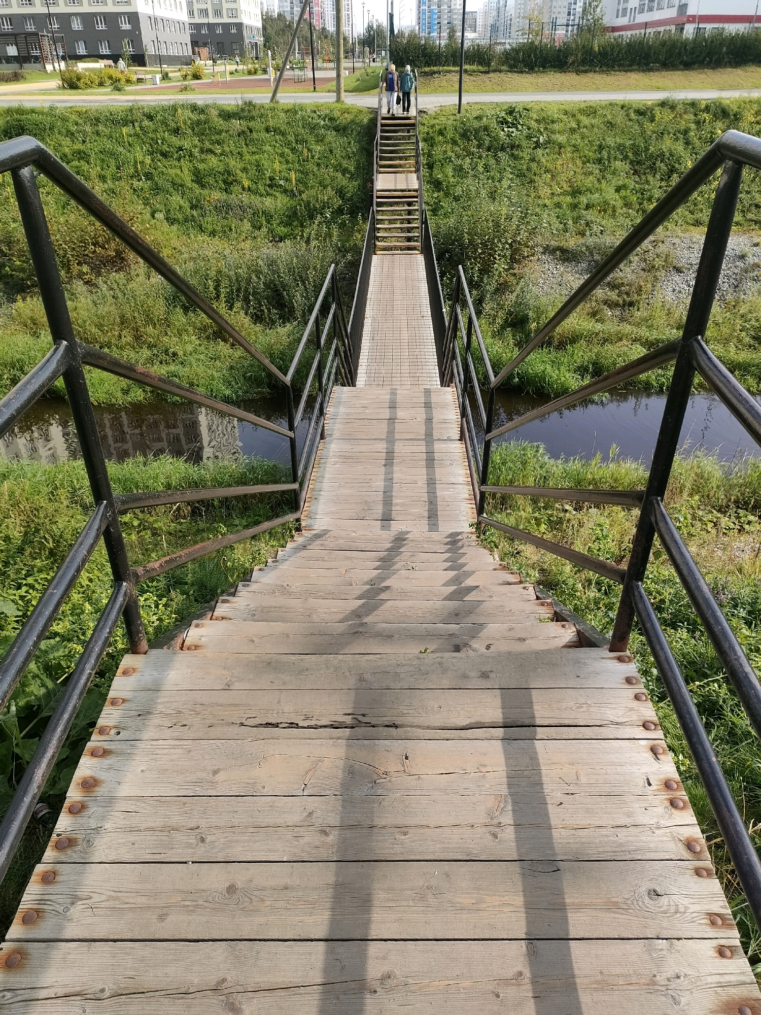 The image shows a wooden pedestrian bridge with metal railings over a small canal or stream. The bridge is made of weathered planks and has stairs leading down towards the viewer. Two people, a woman in a blue jacket and a woman in a green jacket, are walking up the stairs away from the viewer. The bridge is surrounded by green grass and vegetation. In the background, there are modern apartment buildings, a playground, and a road. The scene appears to be in a residential area of Yekaterinburg, Russia, possibly near a park or recreational space. The lighting suggests it is daytime, likely late morning or early afternoon. The overall impression is of an urban landscape integrated with natural elements.