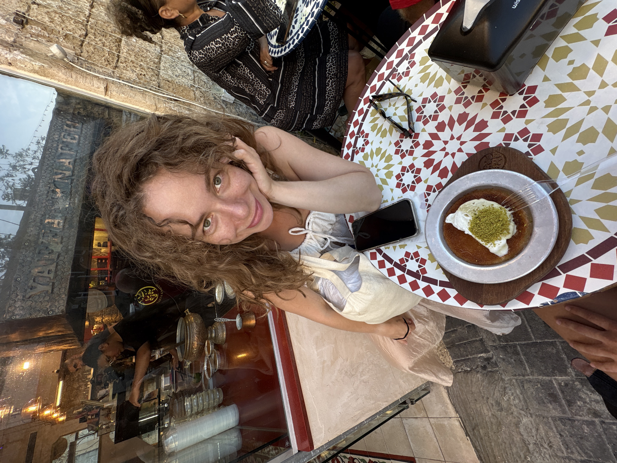 A woman with curly brown hair and green eyes is captured in a selfie, smiling at the camera. She is wearing a white camisole and a light-colored dress, with a tote bag resting on her lap. Next to her, on a table with a colorful geometric pattern, is a dessert topped with whipped cream and chopped pistachios, accompanied by a fork and a smartphone. Sunglasses and a black tissue dispenser are also on the table.
In the background, a glimpse of another woman with dark curly hair wearing a black and white patterned dress can be seen. The setting appears to be an outdoor cafe or restaurant. Reflections in a large glass window reveal a store interior with shelves of items, including what appear to be cups and metalware, and some people working behind a counter. The ground is paved with stone tiles. Visible text on a sign in the window reads "YAFFA". The lighting suggests it is daytime. The woman in the foreground has a relaxed and happy expression.