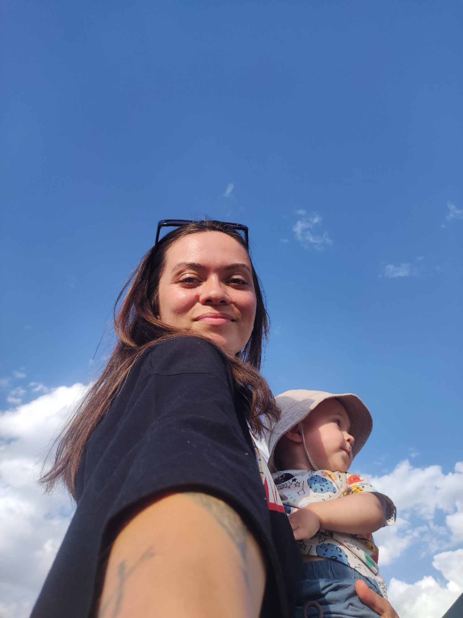 The image depicts a woman holding a young child in an outdoor setting, likely in the city of Chaikovsky, Russia. The woman has long brown hair and is wearing sunglasses on her head and a black t-shirt. She has a tattoo on her upper left arm. The child is wearing a beige sun hat and a patterned shirt. They are both facing toward the upper right of the image. The background consists of a bright blue sky with scattered white clouds.