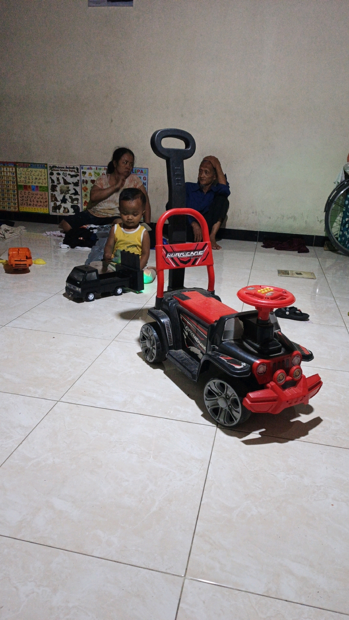 In Bono, Indonesia, a young child wearing a yellow tank top sits on a tiled floor, playing with a black toy truck. To the right of the child is a red and black ride-on toy car with a steering wheel and a push handle. Behind the child, two adults are seated on the floor. A woman wearing a patterned t-shirt and a man in a blue shirt are looking towards the child. Educational posters are displayed on the wall behind them. The environment appears to be an indoor space with cream-colored tiled flooring and a plain wall.