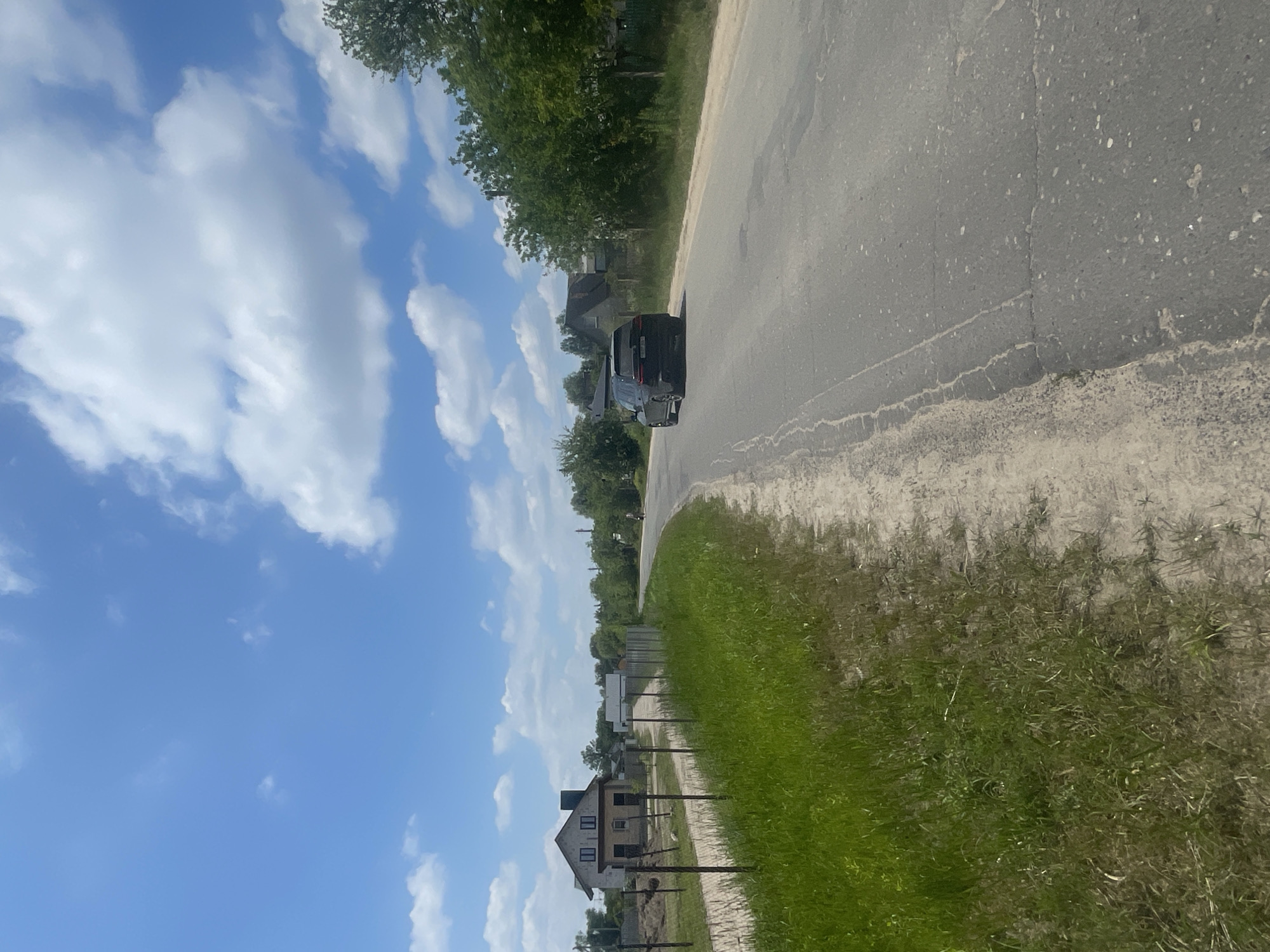 A black SUV is traveling on a paved road in a rural area. The road curves to the right and is bordered by a dirt shoulder and green grass. On the left side of the road, there is a fence and what appears to be a construction site with a partially built house. In the distance, there are more houses and trees. The sky is blue with scattered white clouds. A single person on a bicycle is visible on the road ahead of the SUV. The scene is consistent with a location in or near Мухавецкі сельскі Савет, Belarus.