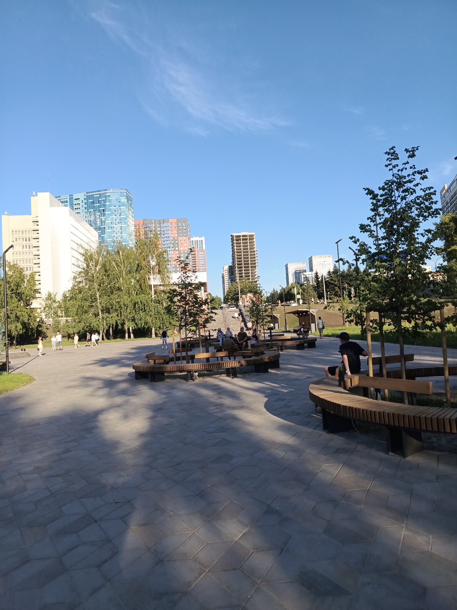 The image captures a modern urban park setting under a clear blue sky. In the background, a cluster of contemporary high-rise buildings dominates the skyline, featuring a mix of architectural styles and colors, including glass facades and visible concrete structures indicating ongoing construction. The foreground is a paved area with an intricate geometric pattern, shaded by scattered trees. Several curved wooden benches are strategically placed throughout the park, some occupied by individuals. One person, wearing a dark t-shirt and shorts, is seated on a bench in the middle right of the frame, facing away from the camera. Other people are visible walking in the mid-ground. Lush green trees and grass add a natural element to the urban landscape. The scene appears to be a public space designed for relaxation and recreation, possibly in the city of Novosibirsk, Russia, given the architectural style of the buildings.