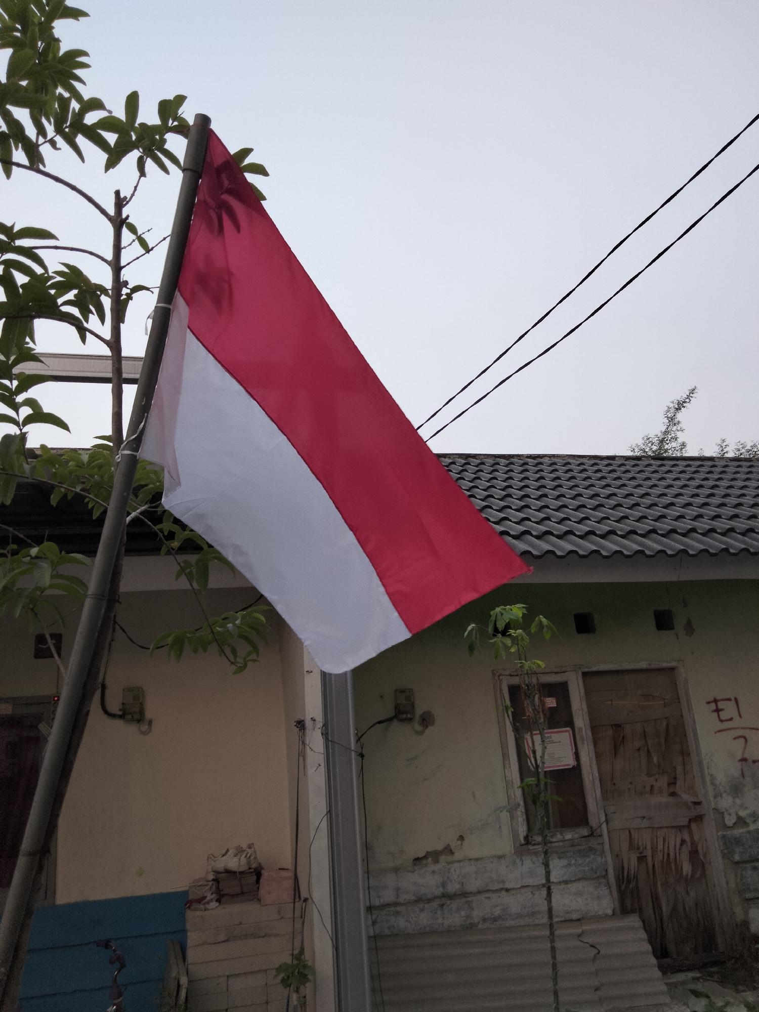 An Indonesian flag, red over white, flies from a flagpole to the left of a residential structure. The building has a dark, tiled roof and pale yellow walls. The door and windows appear weathered. The flag is positioned between a leafy tree and the building. Electrical wires are visible in the sky above the roof. The overall scene suggests an outdoor setting in the town of Cariu, Indonesia.