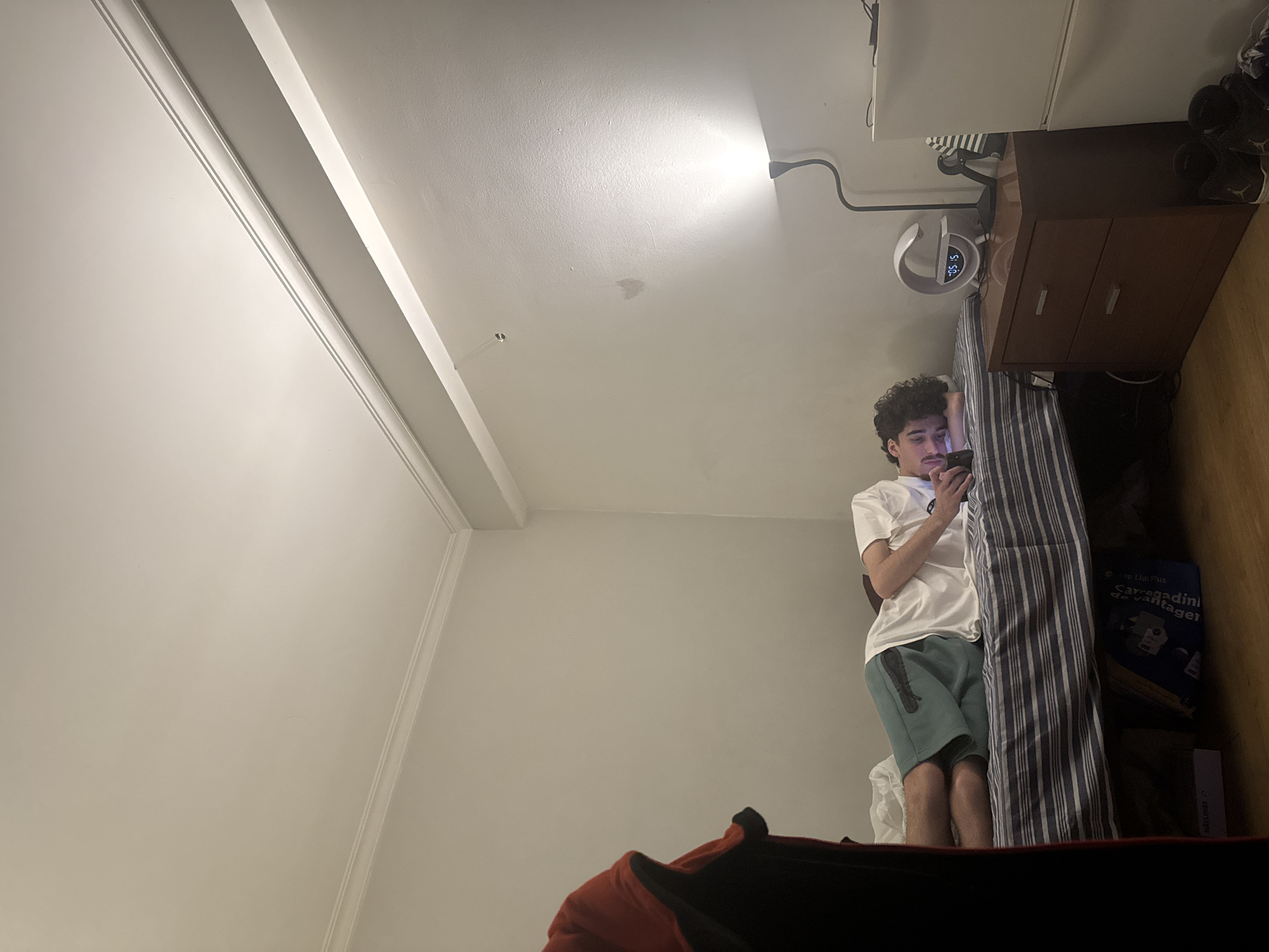 A young man with curly dark hair is standing in a bedroom, looking at his phone. He is wearing a white t-shirt and green shorts. He is leaning against a striped bed frame. To his right is a dark wood nightstand with two drawers and a lamp. On top of the nightstand are shoes. The room has white walls and ceiling with decorative molding. A desk lamp is clipped to the top of the nightstand. The location is likely inside a house or apartment, possibly in Bragança, Portugal, given the presence of the game "Carragadinha de Vantagens" which is a game sold by the Lidl chain of stores in Portugal.