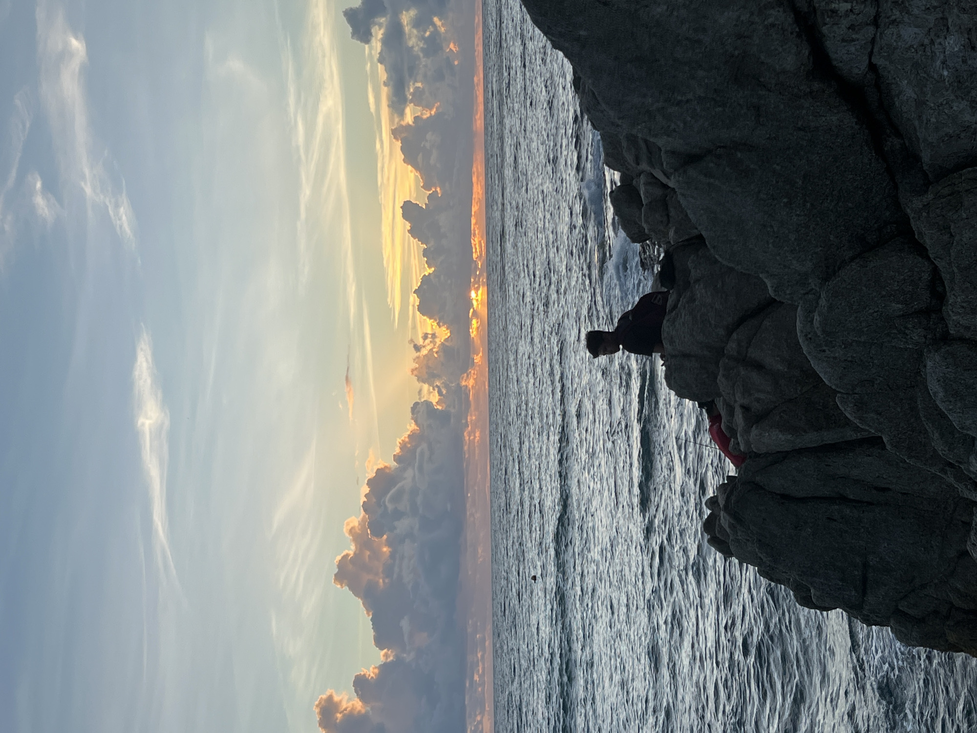 A person sits on a rocky shore overlooking a body of water at sunset. The sky is filled with colorful clouds, illuminated by the setting sun. The water is choppy, with small waves breaking against the rocks. The person is silhouetted against the sky, making it difficult to discern their features. A red object, possibly a backpack or clothing, is near the person. The scene appears to be a natural landscape, possibly a beach or a coastal area, given the presence of rocks and the ocean. The image has a serene and tranquil atmosphere, typical of a sunset scene.