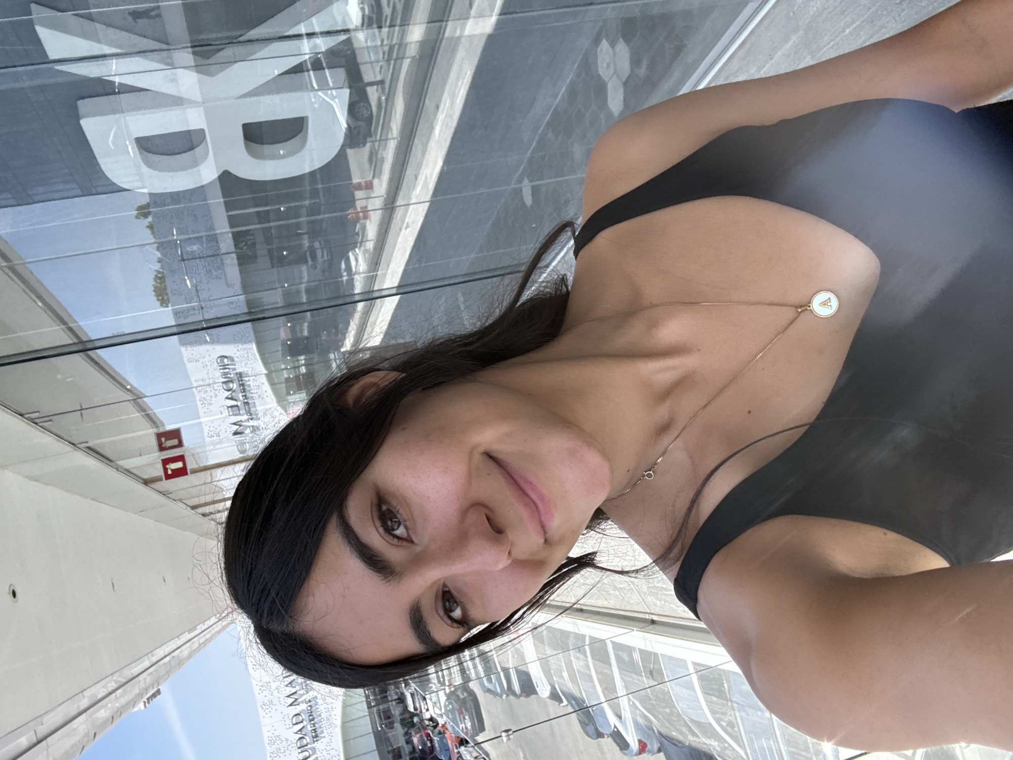 A selfie shows a woman with dark hair and brown eyes wearing a black tank top and a gold necklace with a white and gold pendant. The reflection in the glass behind her shows a large white stylized "B" logo. The reflection also reveals the words "CIUDAD MADRID" and a pedestrian area with cars and people. There's a red sign with two white arrows on the wall. The location is Puebla, Mexico.