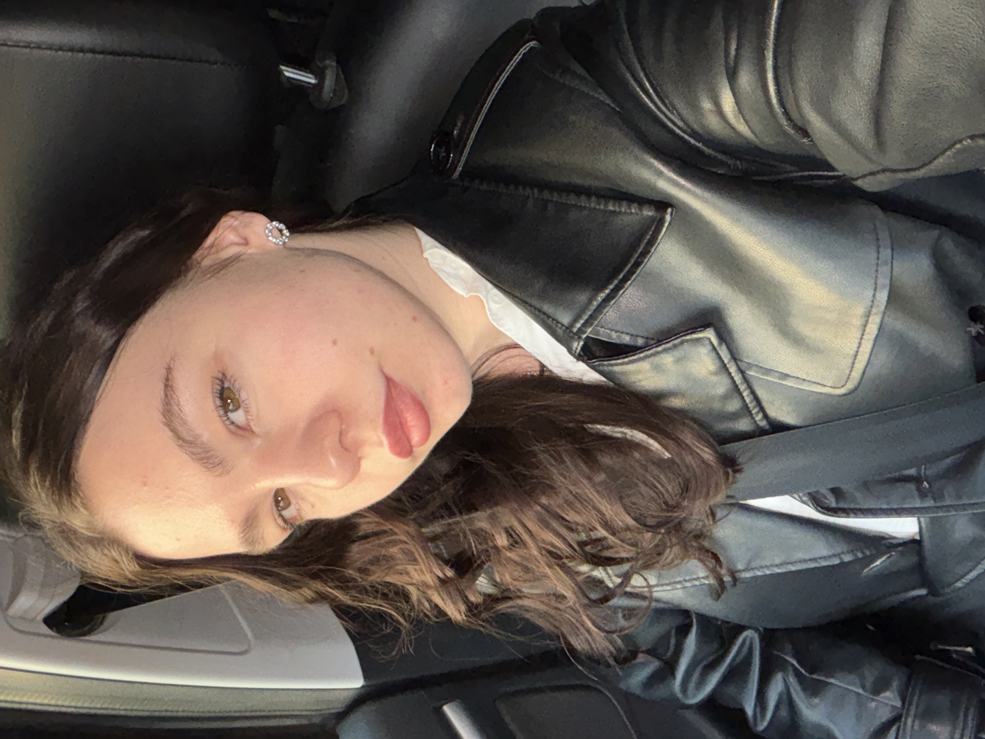 A young woman is sitting in a car, wearing a black leather jacket over a white collared shirt. She has brown hair, brown eyes, and fair skin. She is wearing a round, possibly jeweled earring in her left ear. The car's interior is black and gray. A seatbelt is visible across her torso. The location is not discernable from the image.