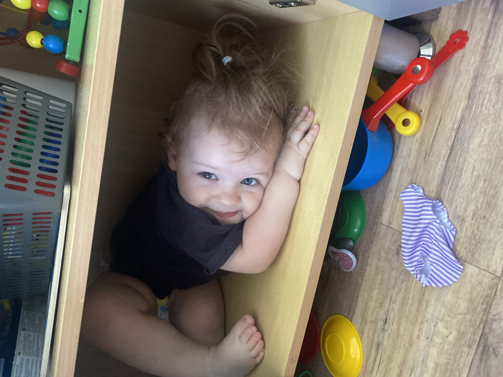 A baby with light brown hair tied in a small ponytail is playfully peeking out from inside an open wooden cabinet or shelf. The baby is wearing a dark-colored top and has bright blue eyes, looking directly at the viewer with a broad, mischievous smile. One arm is propped against the light wood interior of the cabinet, while their legs and feet are curled up beneath them.
The setting appears to be an indoor room with light-colored wooden or laminate flooring. Surrounding the cabinet are various colorful children's toys. To the left, partly visible inside the cabinet or next to it, is a wire maze toy with red, yellow, blue, and green beads, and a gray, gridded plastic container or basket. To the right of the cabinet, on the floor, are several plastic toys including a red spade-like tool, a yellow handle or scoop, a blue bucket, and green, red, and yellow plastic bowls or cups. A small item of purple and white striped clothing, possibly a sock or underwear, is also on the floor. The overall impression is one of a child engaging in playful exploration. There is no visible text in the image.