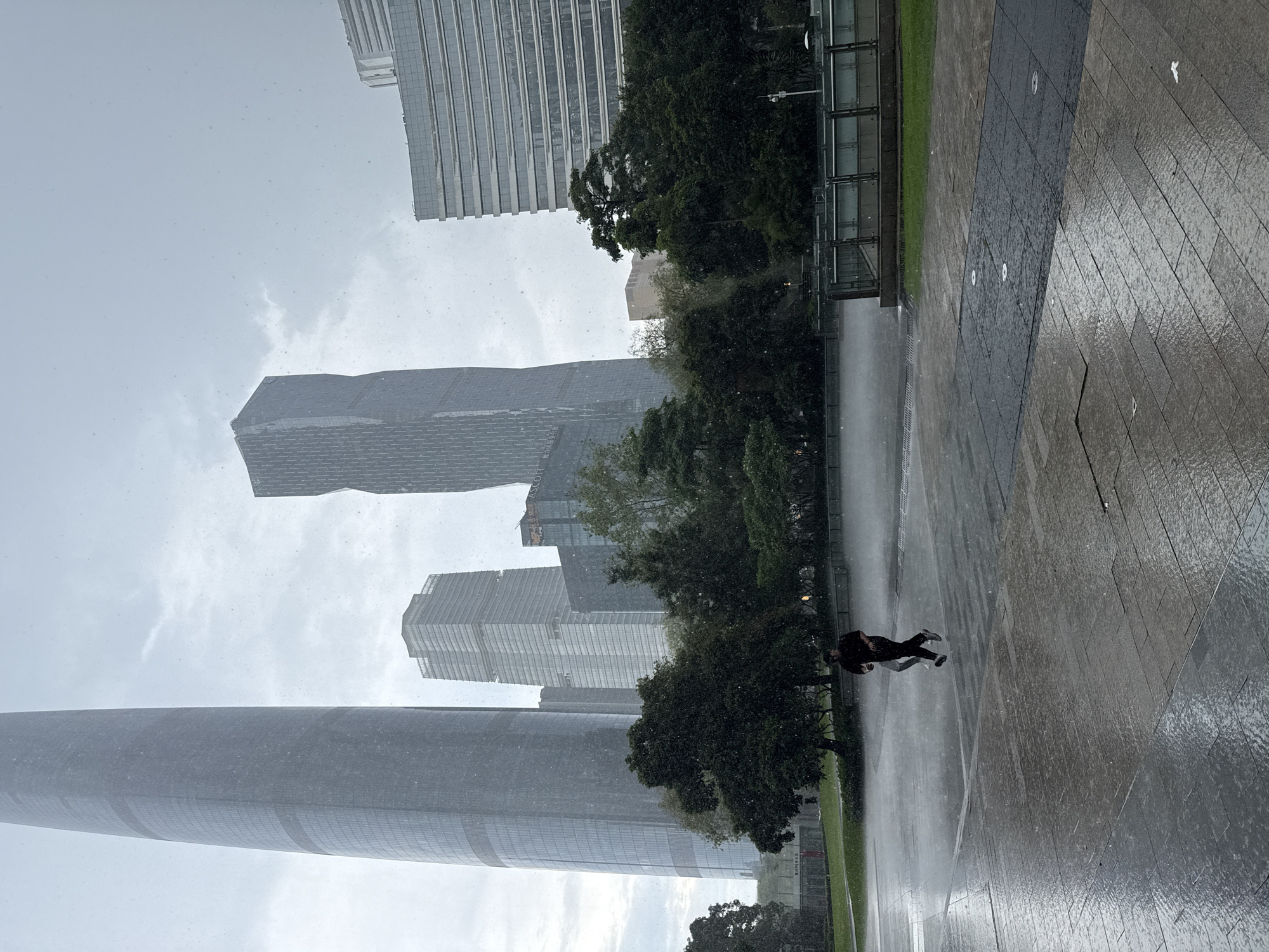 The image depicts a cityscape in Tianhe District, China, during a rain shower. Several modern skyscrapers are visible in the background, including a tall, cylindrical tower on the left and a twisted, angular tower in the center. In the foreground, two individuals are running on a wet, paved plaza. Trees and green spaces are situated between the plaza and the buildings. The rain is visible as streaks and droplets in the air, and the ground is reflecting the ambient light due to wetness. The sky is overcast, contributing to a muted color palette.