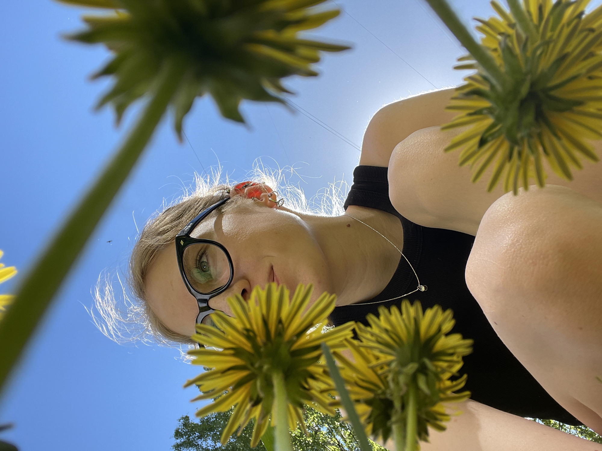A low-angle shot shows a woman with blonde hair and black-rimmed glasses looking down at the camera. She wears a black top and a thin silver necklace with a small charm. Several dandelions are prominently positioned in the foreground and partially obscure the view. The background reveals a clear blue sky and some trees in the distance. The image was likely taken outdoors on a sunny day. The specific location in Izhevsk, Russia, cannot be determined from the image.