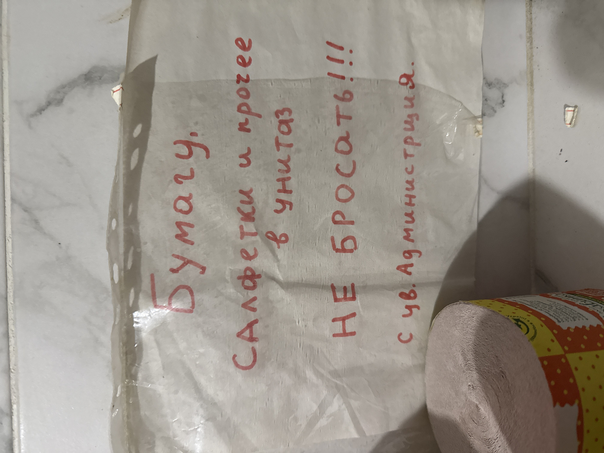 A handwritten note on a transparent plastic sheet is taped to a wall. The note, written in red marker, reads: "Бумагу, салфетки и прочее в унитаз НЕ БРОСАТЬ!!! с ув. Администрация." This translates to "Paper, napkins and other items into the toilet DO NOT THROW!!! Sincerely, Administration." Below the note, a roll of pink toilet paper is visible on the left side of the frame. The wall appears to be tiled with a light-colored marble pattern. The background is a light gray, and the overall lighting suggests an indoor environment, possibly a restroom.