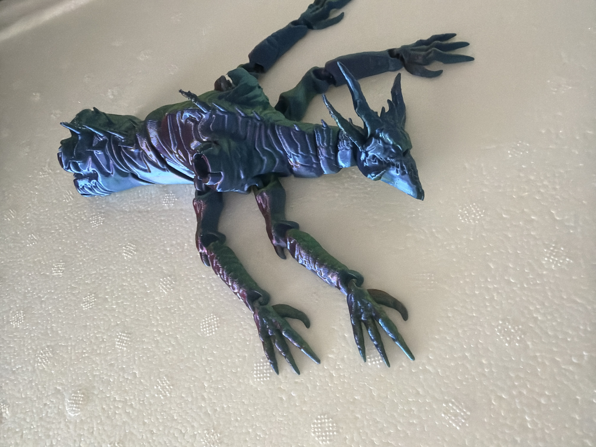 An iridescent, multi-limbed creature figurine is centrally positioned on a white, textured surface. The figurine exhibits a shimmering color palette, primarily deep blue transitioning to teal and hints of purple, suggesting a metallic or pearlescent finish. It possesses a distinct head featuring prominent spikes and a pointed snout. The body is characterized by a segmented or ridged texture, indicative of scales or armor. Four distinct forelimbs, each terminating in multi-clawed hands, extend from the upper torso. Two hind limbs, also ending in clawed feet, are visible towards the lower body. The figurine is oriented on its back, with its head directed towards the bottom-right of the frame and its body extending diagonally upwards to the left. The limbs are splayed outwards. The underlying surface is uniformly white and features a repetitive pattern of small, raised, square-grid depressions, resembling a padded or quilted material. The scene is evenly illuminated, casting minimal shadows. The location is Unknown, Unknown.