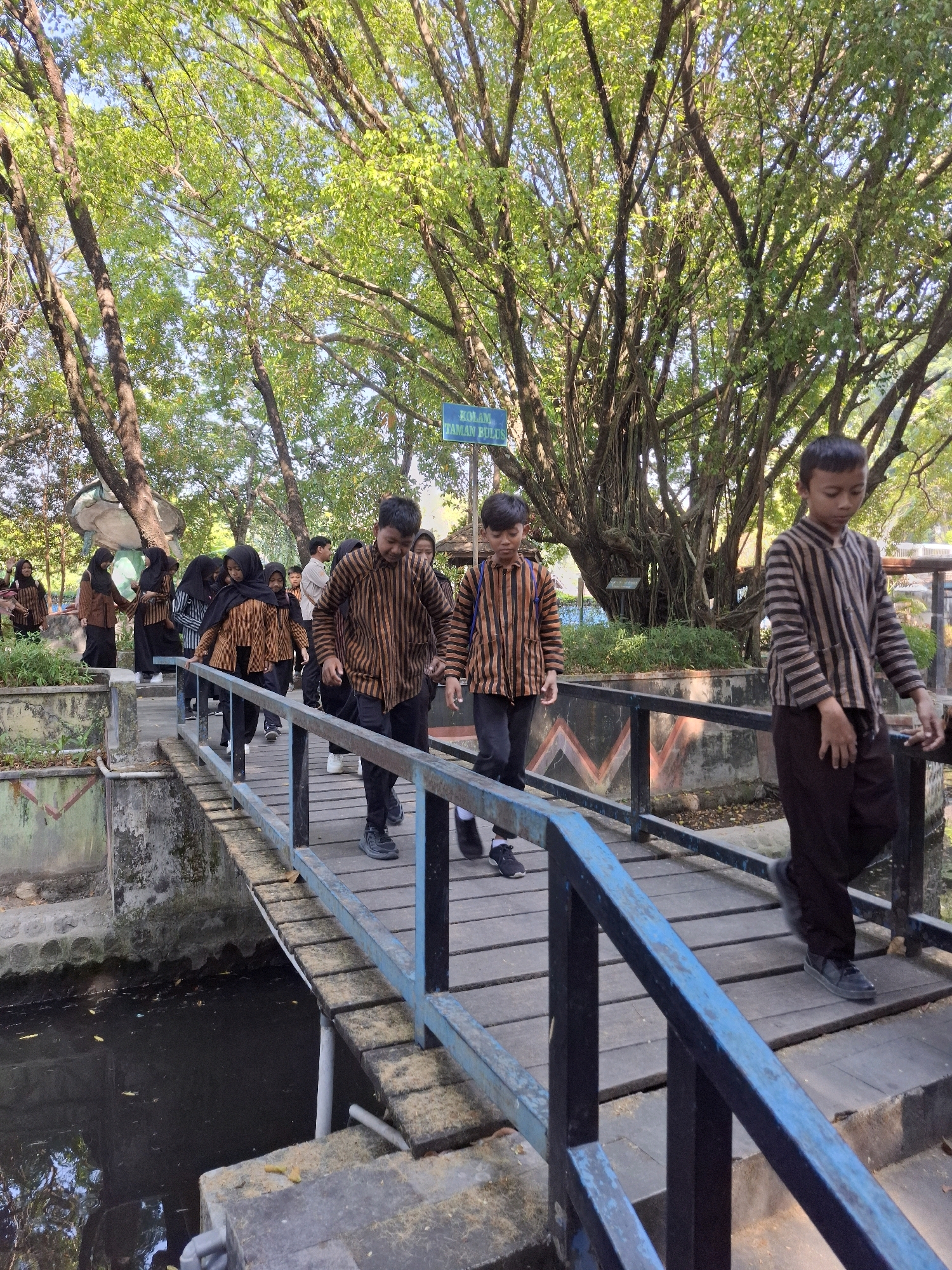 In Tawun, Indonesia, a group of children, likely students, are walking across a wooden bridge. The children are dressed in matching striped shirts with brown and tan patterns, and some wear headscarves. They are walking in the same direction, suggesting they are part of a group excursion or school activity. The bridge is weathered and has blue and black railings. Below the bridge is a dark body of water. The surrounding environment is lush with trees, their leaves forming a canopy overhead, and a bright sky is visible through the branches. In the background, there are concrete structures and more greenery, indicating an outdoor park or educational facility. The sun is shining, casting dappled light through the trees onto the bridge and the children. A sign is visible in the upper left of the image, partially obscured by trees, indicating this is a public area.