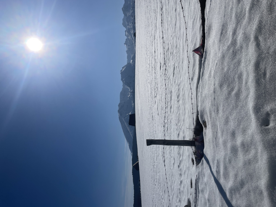 The scene depicts a snow-covered winter landscape in Palež, Montenegro, under a clear, bright blue sky. In the foreground, a wooden fence post stands upright, partially embedded in the snow. Two strands of barbed wire extend horizontally from the post across the lower portion of the image. To the right of the post, a red and white object, possibly a piece of plastic or fabric, is partially buried in the snow. A distinct shadow from the fence post stretches towards the lower left. The midground features an expansive, flat field uniformly covered in snow, with two dark-roofed structures, likely buildings, visible on the left and center-right, appearing low against the snowy horizon. In the background, a range of prominent snow-capped mountains rises. The sun is visible in the upper right quadrant of the sky, emitting bright rays and lens flare, indicating a clear day with strong overhead sunlight. No individuals are present.