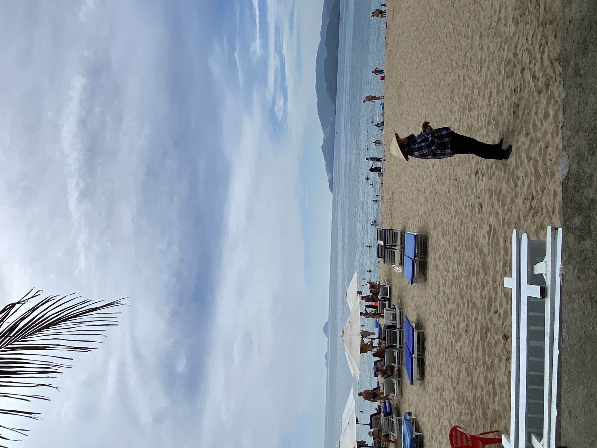 This image depicts a crowded beach scene in Nha Trang, Vietnam, likely on a cloudy day, as evidenced by the overcast sky and muted lighting. In the foreground, a person wearing a conical hat and a plaid shirt stands on the sand, facing away from the camera and toward the ocean. The beach is lined with numerous lounge chairs and umbrellas, some occupied by beachgoers. The ocean is calm with visible waves, and several people are swimming or wading in the water. In the distance, hills and mountains form the coastline. A palm frond is visible in the upper left corner, adding a tropical element to the scene. The presence of lounge chairs, umbrellas, and a diverse group of people suggests a popular tourist destination within Nha Trang.