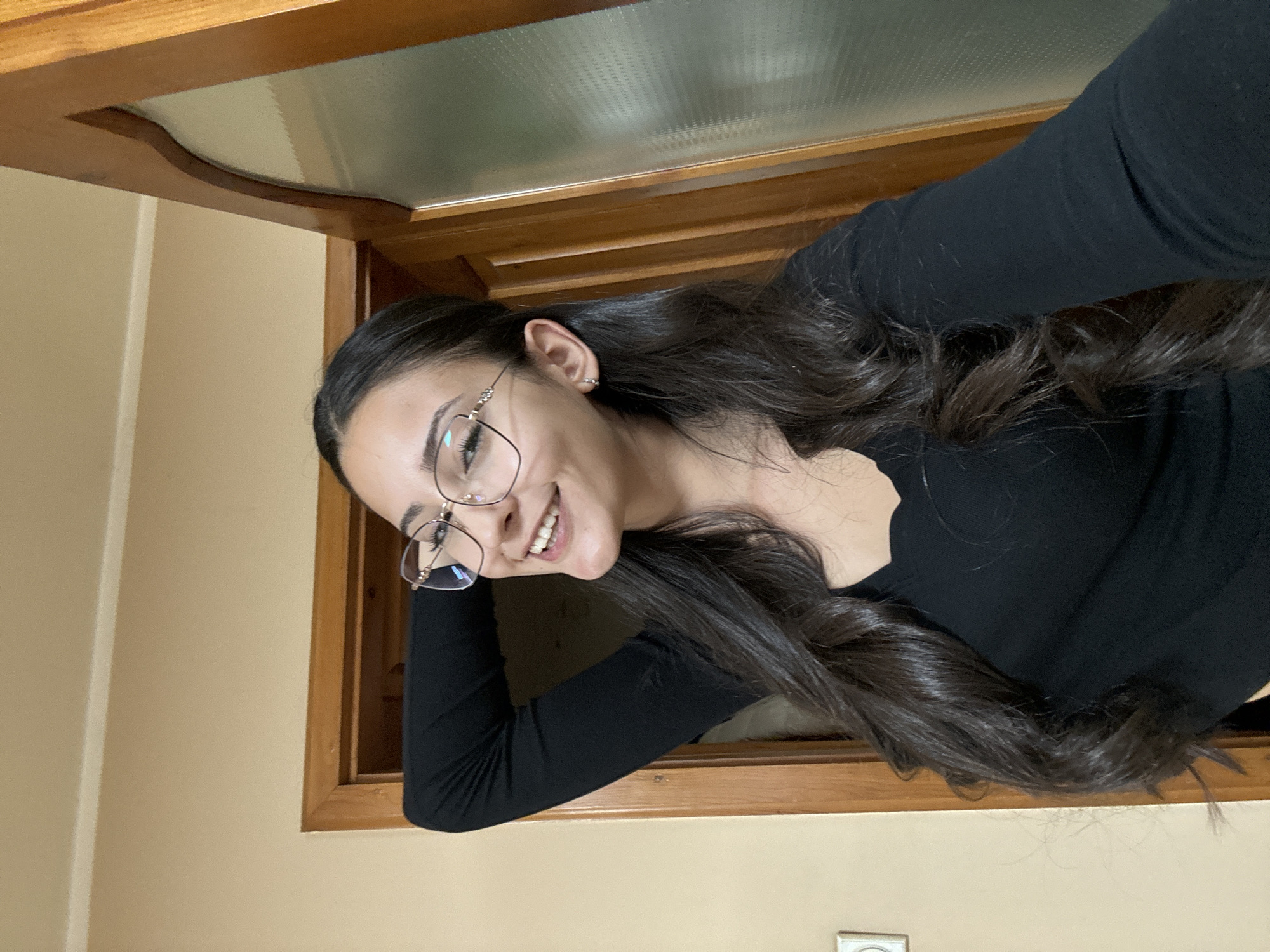 A brunette woman wearing glasses and a black long-sleeved shirt is leaning against a wooden-framed window or doorway. She is smiling and looking towards the camera. The location appears to be indoors, with beige walls. The city of Dupnitsa, Bulgaria is not discernible from the image.