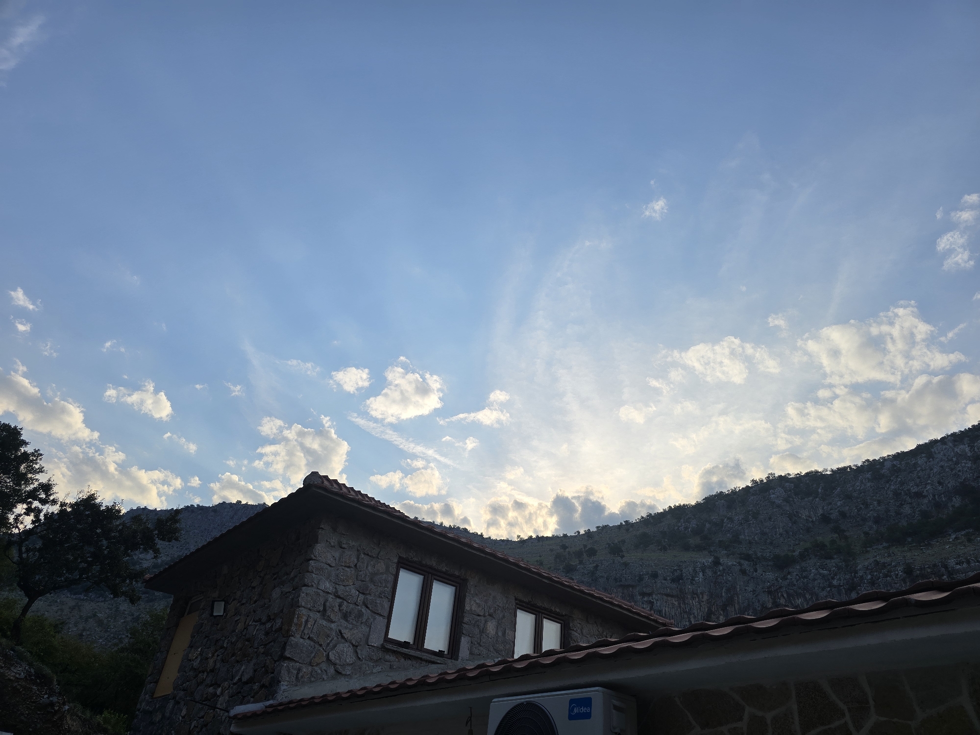 The image captures a house made of stone with a red tiled roof, partially silhouetted against a dramatic sky filled with wispy clouds and patches of sunlight. The house has two windows with dark frames. An air conditioning unit is mounted on the exterior wall below the roofline. Behind the house, a steep, rocky mountainside rises, covered in sparse vegetation. To the left of the house, a large tree with dark foliage is visible. The overall scene suggests a rural or mountainous setting. The location is identified as Podgorica, Montenegro.