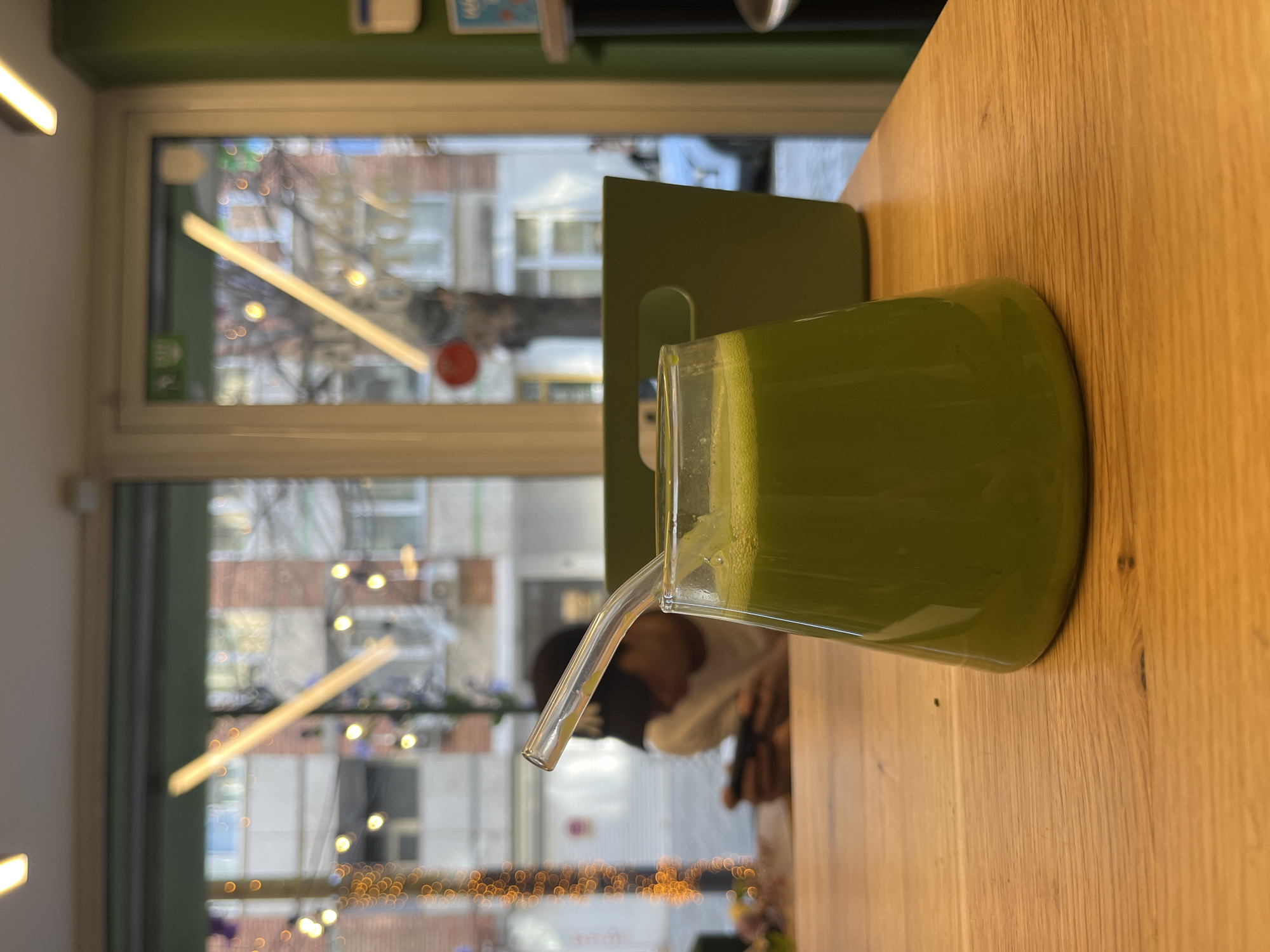 A glass of green juice with a clear straw sits on a wooden table. Behind the glass is a green vertical object. Through a window, a view of a city street with buildings, decorative lights, and a person visible in the background. The location is Sofia, Bulgaria.