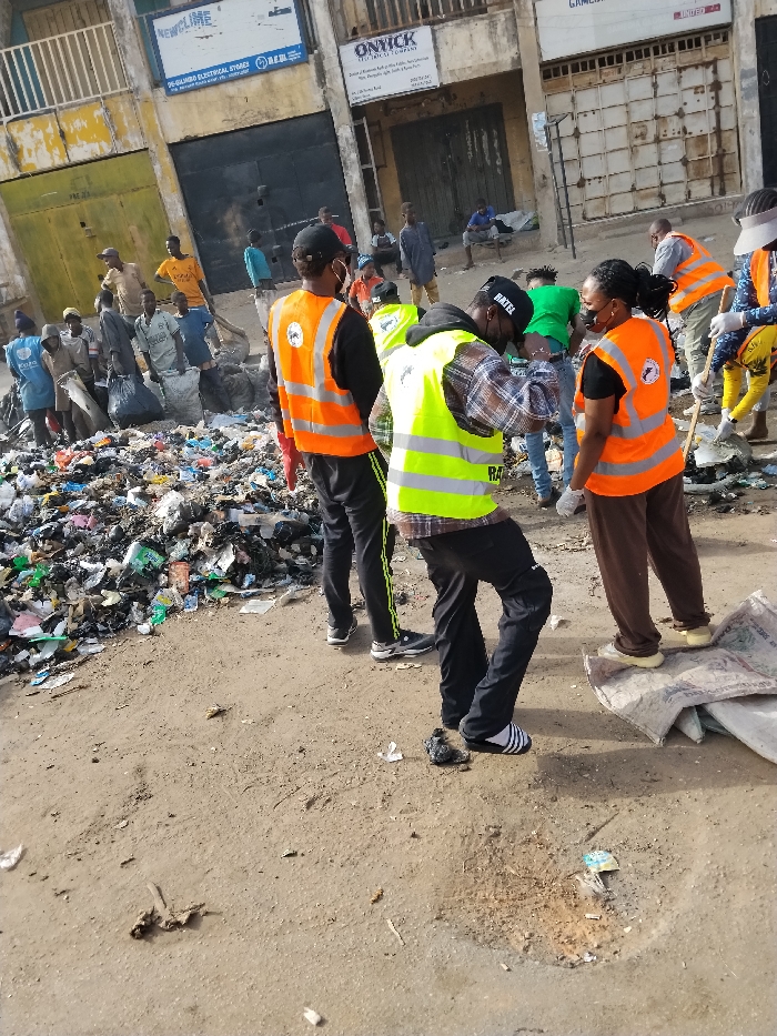 The image displays a daylight scene on a dusty, unpaved street in Fagge C, Kano State, Nigeria, specifically along Kano Street as indicated by signage. A large, sprawling accumulation of mixed refuse, including plastic bags, fabric, and various debris, occupies the left side of the midground. Multiple individuals are present, many actively engaged in or observing a cleanup operation.
In the foreground and midground, several individuals are prominent. A man wearing a neon green high-visibility vest, a black cap with "RATEL" visible, black trousers, and striped sandals is bending forward. To his immediate left, another man in an orange high-visibility vest, black shirt, and black track pants with green stripes stands with his back partially to the camera. To the right of the man in the green vest, a woman wearing an orange high-visibility vest, a black shirt, brown trousers, and a black face mask stands, facing the man in the green vest.
In the background, additional individuals are clustered around the refuse pile, some also wearing orange high-visibility vests and seemingly handling waste, with one individual on the far right holding a tool resembling a shovel or broom. Other individuals are visible in the distance, some standing, one seated on a low structure. The background consists of multi-story buildings with partially obscured or closed ground-floor storefronts. Visible signage includes "NEWCLIME / 90-GOMBO ELECTRICAL STORE" and "ONYICK INDUSTRIAL COMPANY / ... Kano Street, Fagge, Kano State." The scene depicts a community cleanup effort.