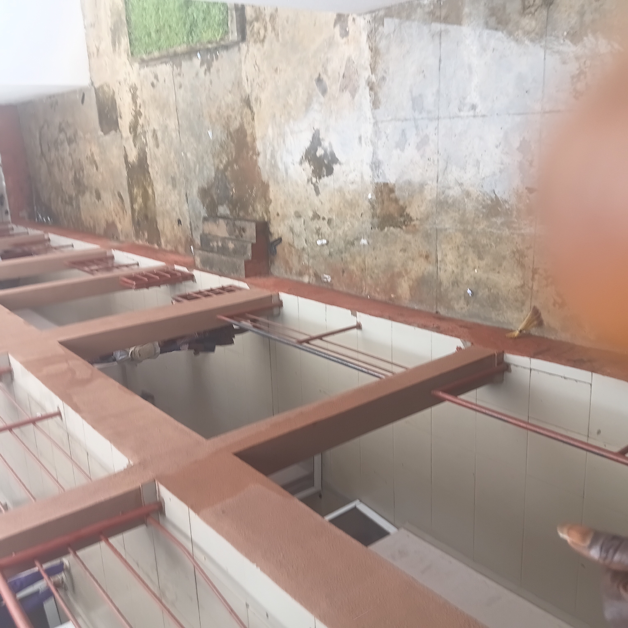 The media file depicts an outdoor scene in Wukari, Nigeria, likely an area designed for laundry or general utility. The setting features a series of reddish-brown metal structures, possibly drying racks or shelving units, arranged in a grid-like pattern. These structures are suspended over a tiled floor, suggesting an indoor or semi-enclosed space.
The background shows a weathered concrete wall with patches of green vegetation visible at the top left, hinting at an exterior wall or boundary. The wall's condition suggests it might be an older structure, possibly exposed to the elements. There are some debris or stains on the wall, and a broom leaning against it on the right side of the image, indicating recent activity or maintenance.
The lighting suggests it is daytime, with no specific indicators of time of day beyond natural light. The presence of what appear to be clothes hanging on some of the racks implies that laundry is being done or has been done. A blurry human finger is visible in the lower right corner of the image, suggesting someone is taking the photograph or is present in the scene, but their face or the rest of their body is not clearly visible.
Overall, the scene conveys a sense of functionality and perhaps a utilitarian aspect of daily life in the depicted location. There is no visible text in the image.