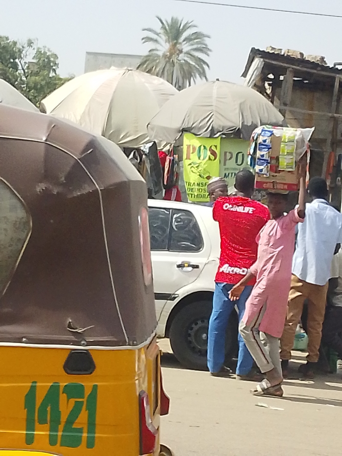 This image captures a street scene in Fagge A, Nigeria. In the foreground, the rear section of a yellow and brown three-wheeled auto-rickshaw is visible, bearing the green numbers "1421". To its right, a white four-door sedan is parked or stationary. Several individuals are present: a person in a red shirt displaying "OGUNLIFE" and "AKRO" on the back, wearing blue pants and sandals, stands near the white car, facing slightly right. To their left, another person, dressed in a pink tunic over grey striped pants and sandals, carries a cardboard box laden with small packaged goods on their head, facing left. Further right, a man in a light blue shirt and tan pants is seen from behind. Behind the people and vehicles, two large open market umbrellas, one light beige and one grey, provide shade over market stalls. These stalls display hanging clothing and two bright yellow banners with "POS" prominently printed, indicating "TRANSFER, DEPOSITS, WITHDRAWAL" services. A dilapidated structure with a corrugated roof is visible behind the stalls. In the background, a palm tree and other sparse vegetation are present under a bright, hazy sky.
