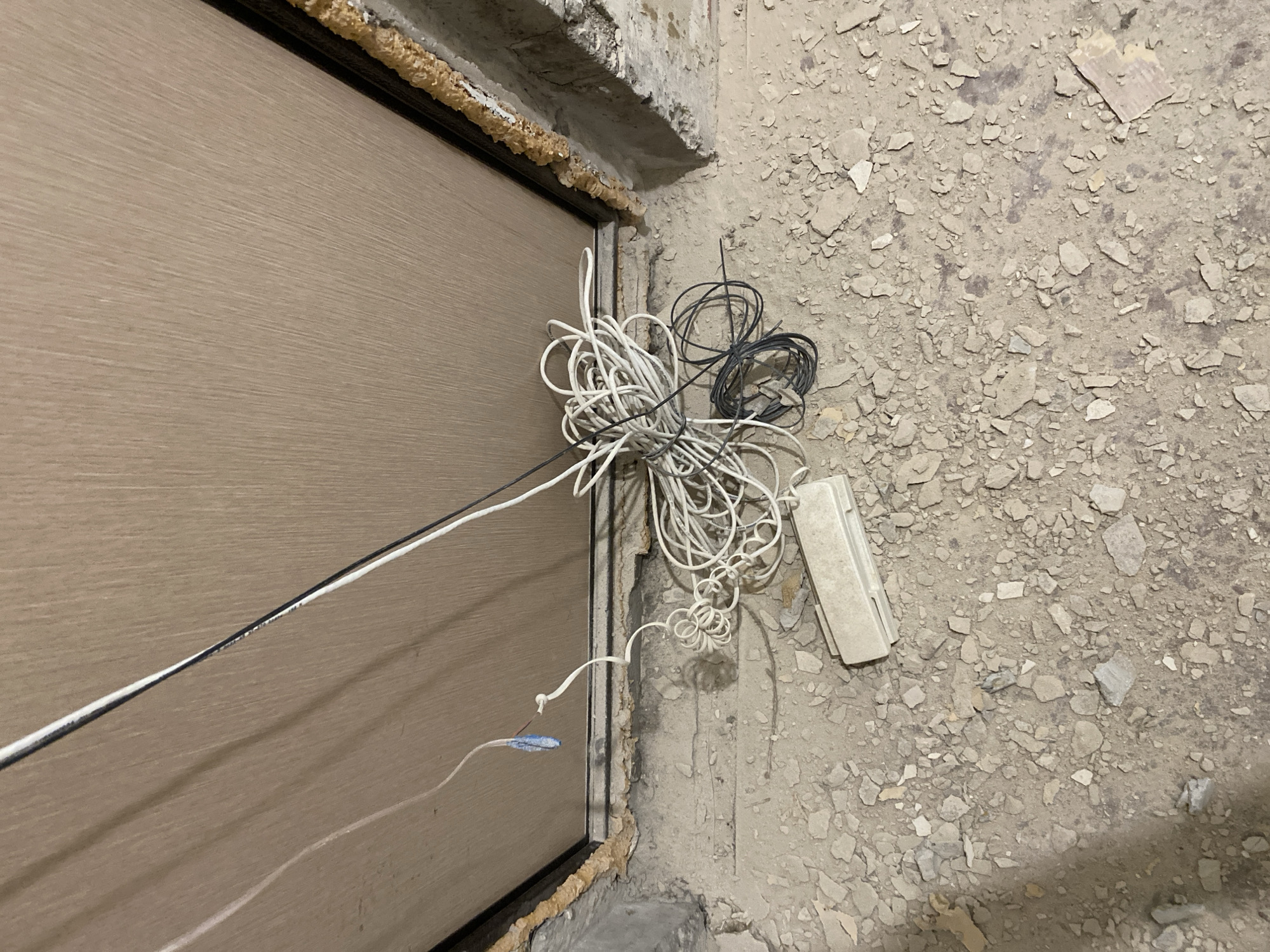This media file depicts a close-up, eye-level view of construction debris and electrical wiring next to a door frame. The scene is set outdoors or in an unfinished indoor space, likely during daylight hours, as indicated by the lighting. The ground is covered in rubble and concrete fragments, suggesting an active construction or demolition site.
The primary objects in the frame are several tangled bundles of electrical wires, one bundle predominantly white and another black. These wires are loosely coiled and appear to be either recently installed or discarded. A white plastic electrical box is visible lying on the rubble. Adjacent to the wires and rubble is the edge of a light brown door, which has a visible seam running down its center. The door frame itself is lined with expanding foam insulation, a common material used in construction to seal gaps.
The overall impression is one of ongoing work, with materials and tools laid out or abandoned. There are no people visible in the image. The location context of Ulyanovsk, Russia, suggests this is a scene from that region. The image is factual and devoid of any discernible text. The focus is on the raw, unfinished state of the area and the presence of electrical components.