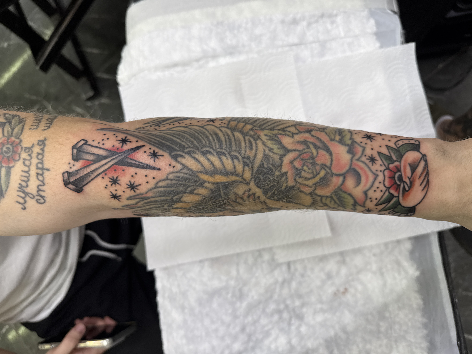 The image shows a close-up of a forearm with multiple traditional-style tattoos. Tattoos visible include two nails crossed over with drops of red ink, a bird with spread wings, a rose, a pair of hands holding each other, and script in Cyrillic (possibly Russian). The arm is resting on white paper towels. Part of a chair and a phone can be seen in the background. The location is not discernible.